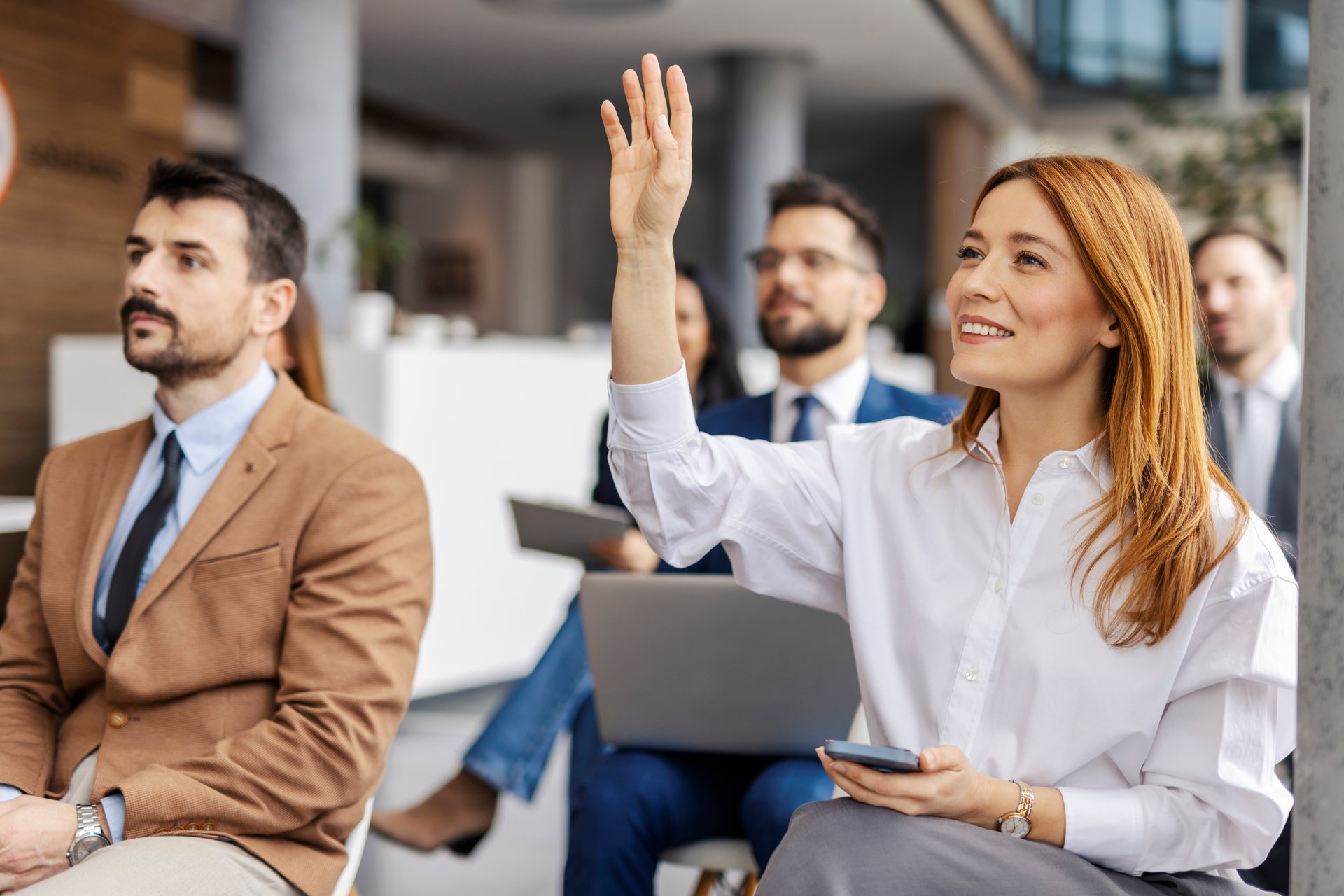 Portrait of smiling female manager sitting with her team at boardroom during seminar, rising hand and asking questions.