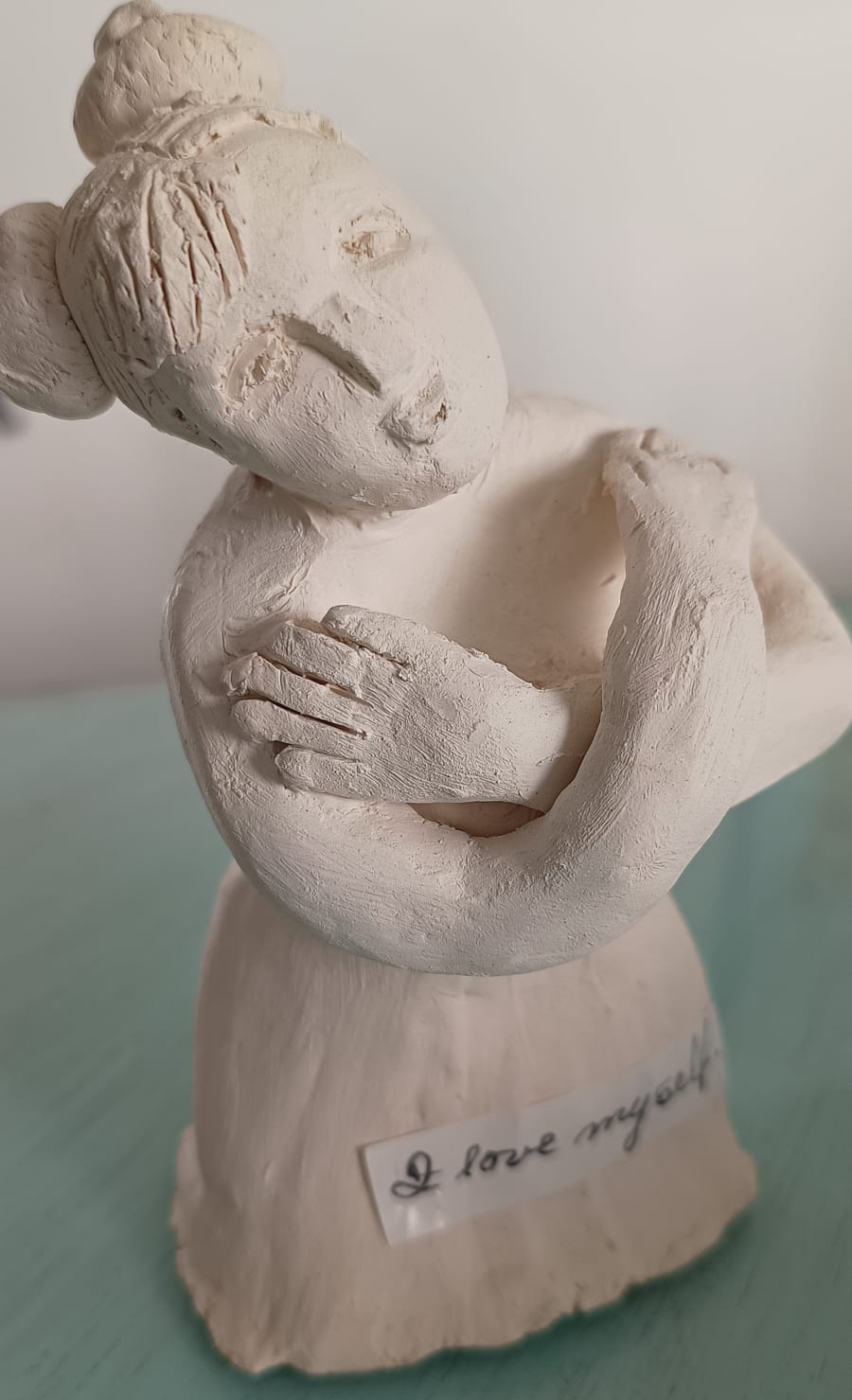 Clay sculpture of a person embracing themselves, with a small tag reading I love myself on the base.