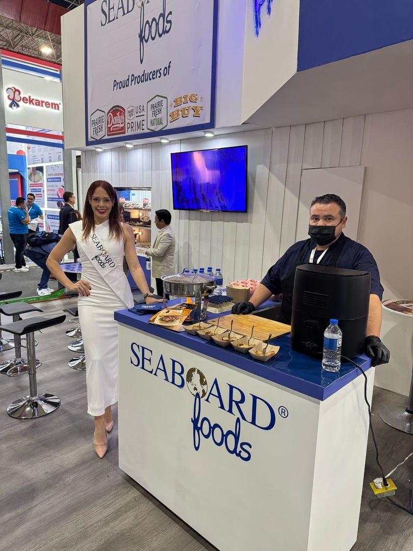 A woman in a white dress stands by a Seaboard Foods booth with a man in a black apron serving food at a trade show.