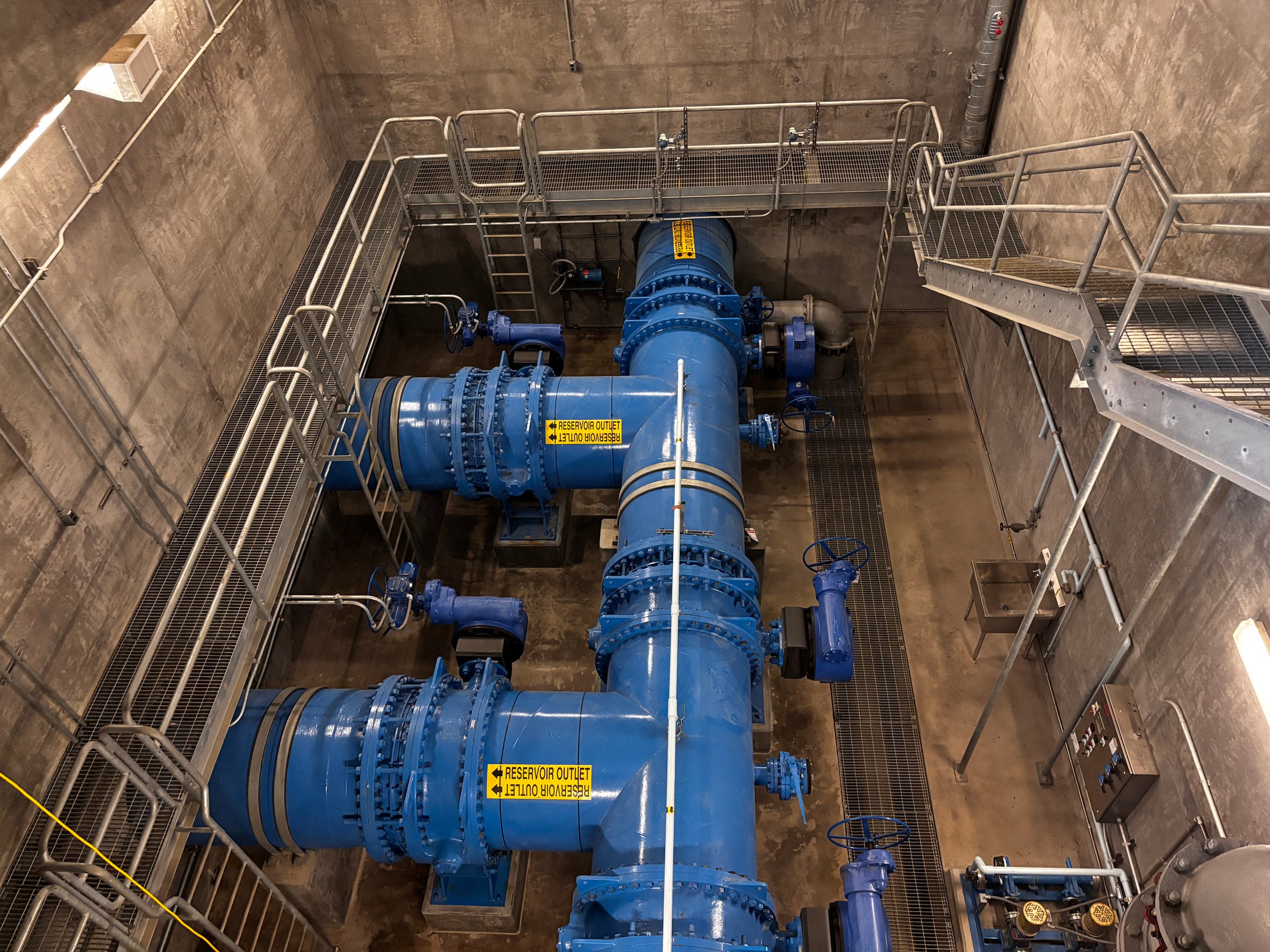 An interior view of a reservoir outlet structure featuring large-diameter pipes, valves, and supporting infrastructure within a reinforced concrete chamber.