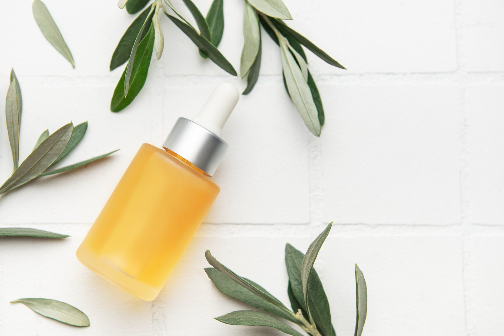 Elegant cosmetic serum bottle with pipette dispenser lying on white tile background decorated with olive tree leaves, promoting natural organic skincare