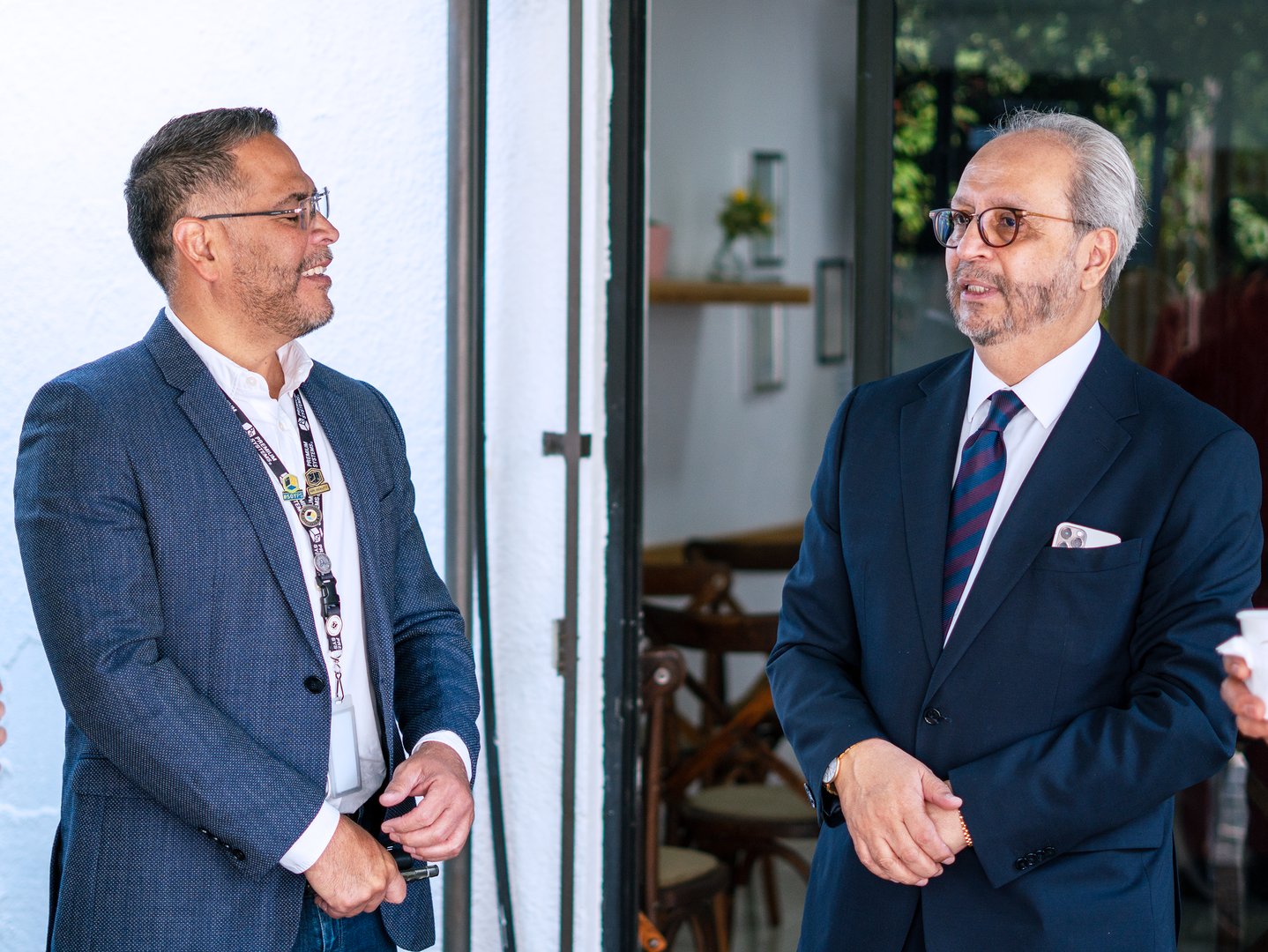Two men in suits smiling and conversing indoors near a door, with one holding a cup. They appear to be at a formal event.