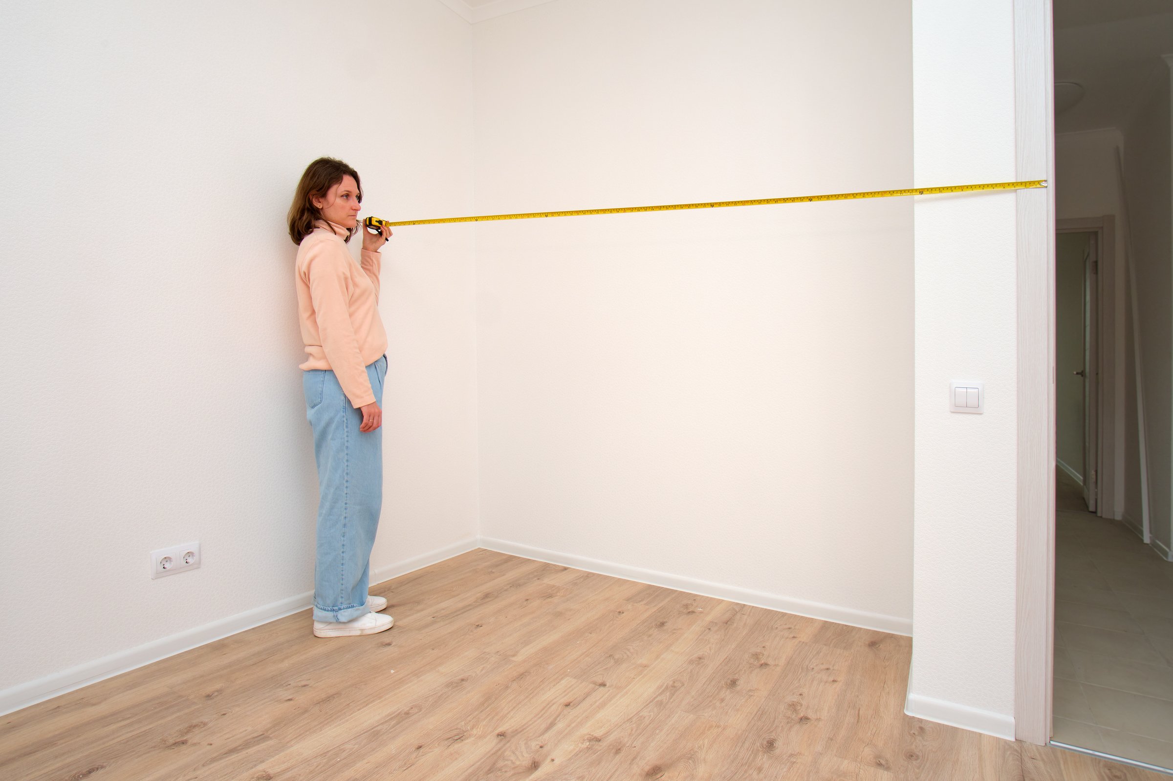 Woman measuring empty room corner with tape measure. Home renovation planning in modern white interior.