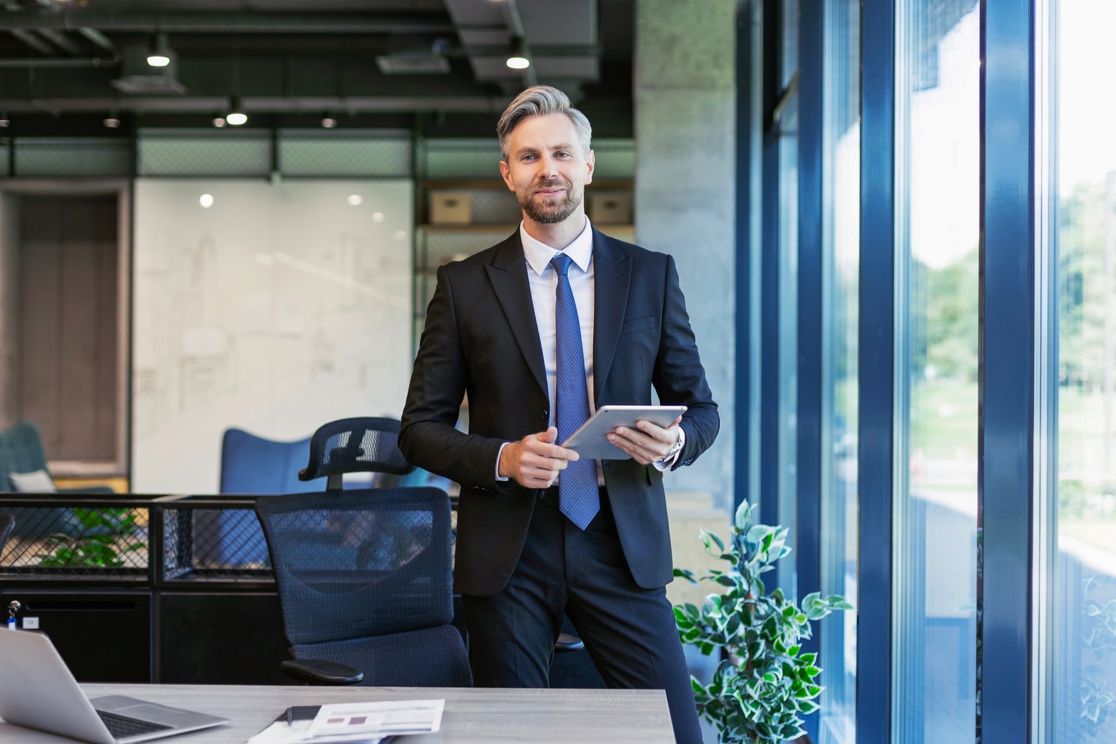 Portrait of smiling middle aged businessman standing with digital tablet at corporate office