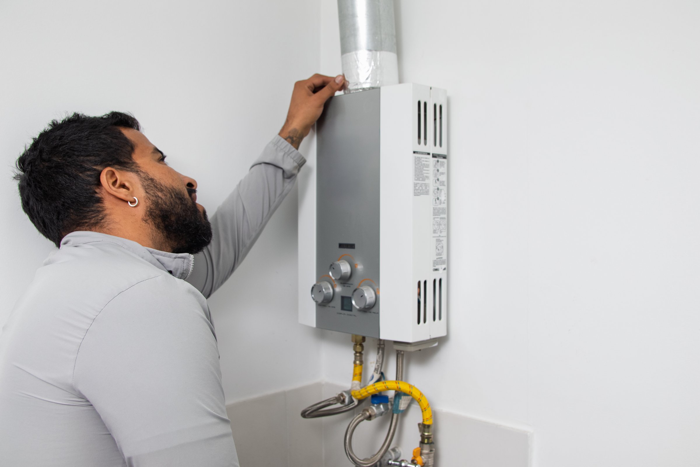 A bearded man in a light grey shirt reaches up to touch or inspect the venting pipe of a wall-mounted, tankless water heater or boiler