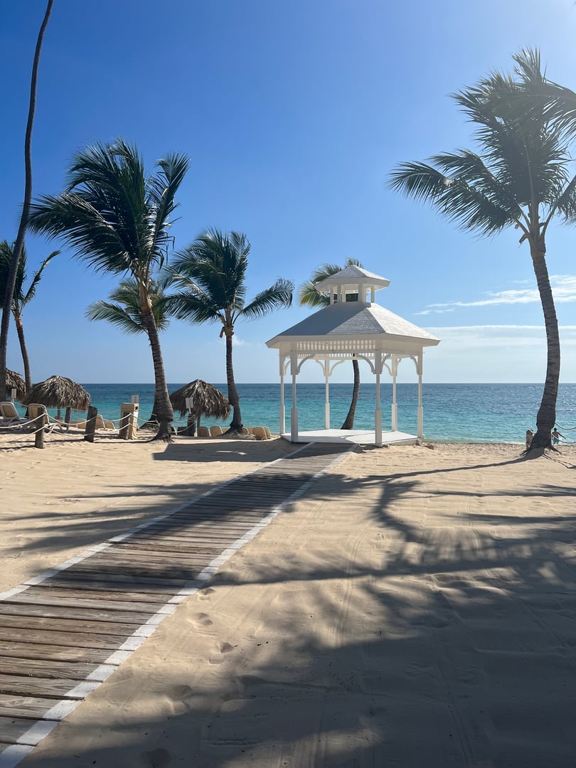 Beach scene with a white gazebo, palm trees, sandy path, and clear blue sky by the ocean.