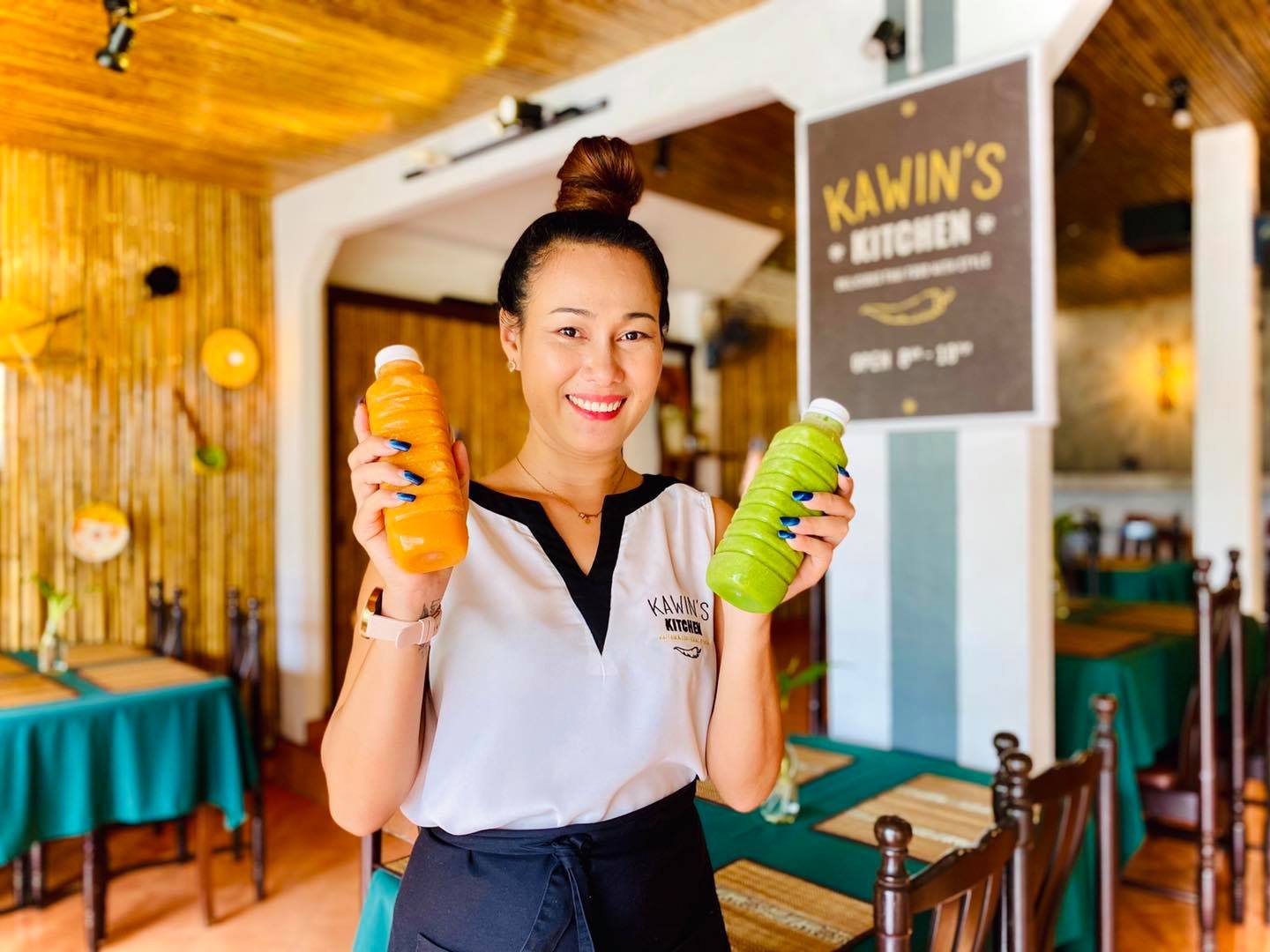 Smiling woman in a restaurant holding a bottle of orange juice and a bottle of green juice.