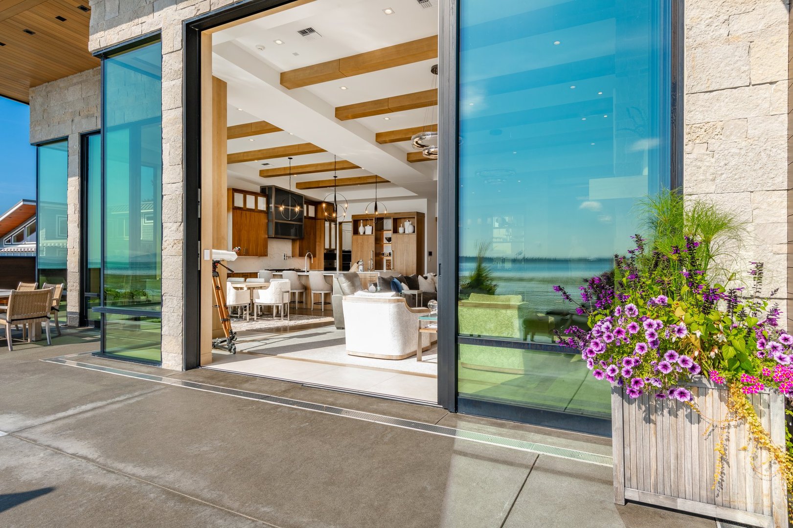 Exterior photograph of a mountain modern home residence a bright blue sky summer day and the house has an expansive ocean front deck with a swimming pool and a hot tub the tide is low and there are coastal views we also see an enclosed outdoor living area with stainless steel cabinets cooking facilities dining area wooden furniture the front facade of the home shows a flat roof and two large garage doors with glass windows the home has stone and wood cladding waterfront deck leads to modern living room through floor to ceiling glass doors