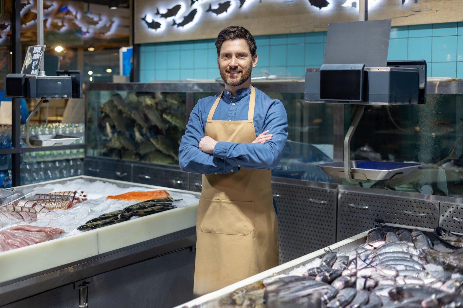 Confident fishmonger standing with arms crossed in front of seafood display at fish market. Fresh fish and seafood in foreground. Man wearing apron smiling at camera.