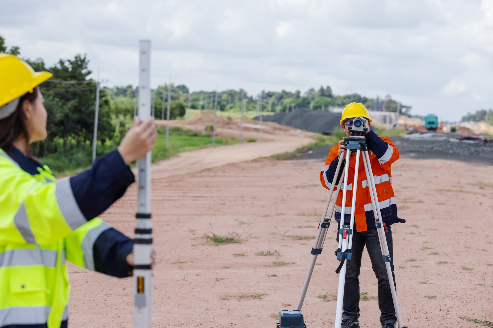 Two survey engineers wearing helmets and reflective jackets work together at a construction site. One holds a leveling staff while the other operates the tripod instrument for land measurement accuracy.