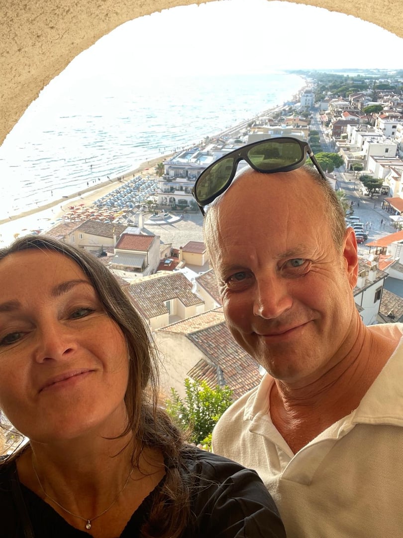 A smiling couple takes a selfie from a high vantage point, overlooking a coastal town and beach with umbrellas and buildings.