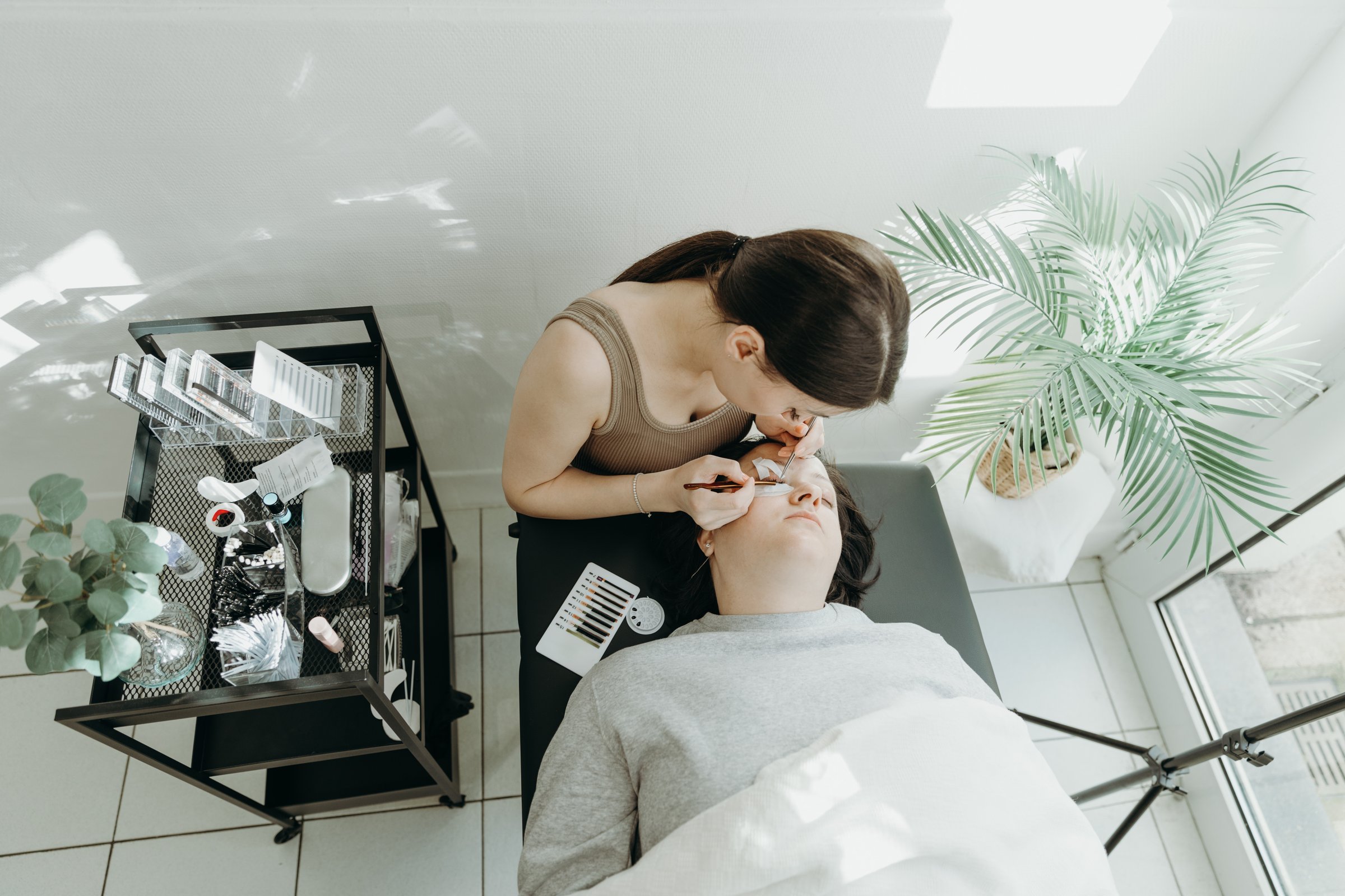 Portrait of one young beautiful Caucasian cosmetologist girl who carefully glues artificial eyelashes on the right eye with the help of two tweezers to a female client lying on the cosmetology table in her home office on a spring day, top view close-up. Concept of eyelash extensions, beauty industry.