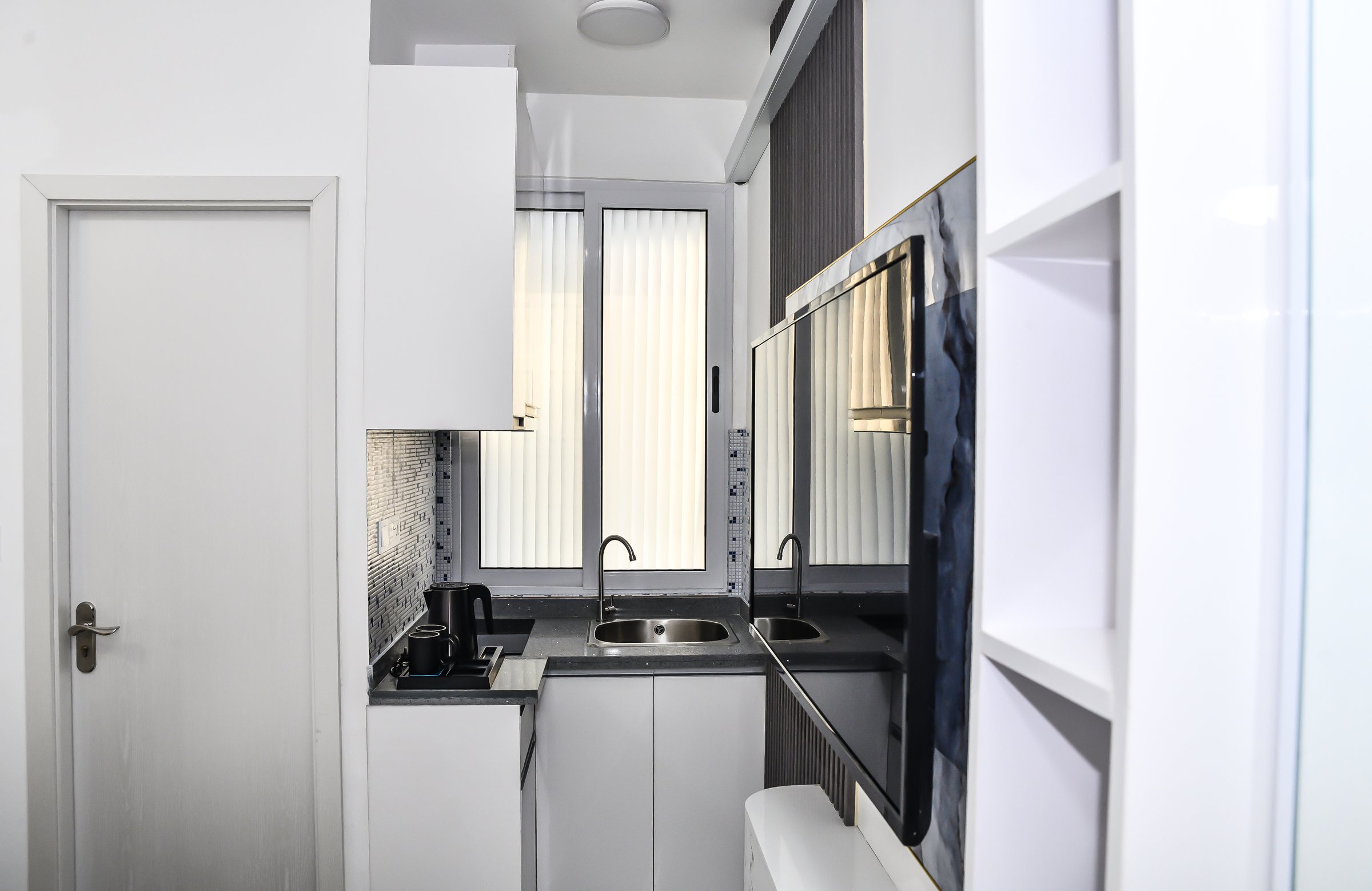 Modern compact kitchen with white cabinets, dual sinks, built-in appliances, and a mosaic tile backsplash.