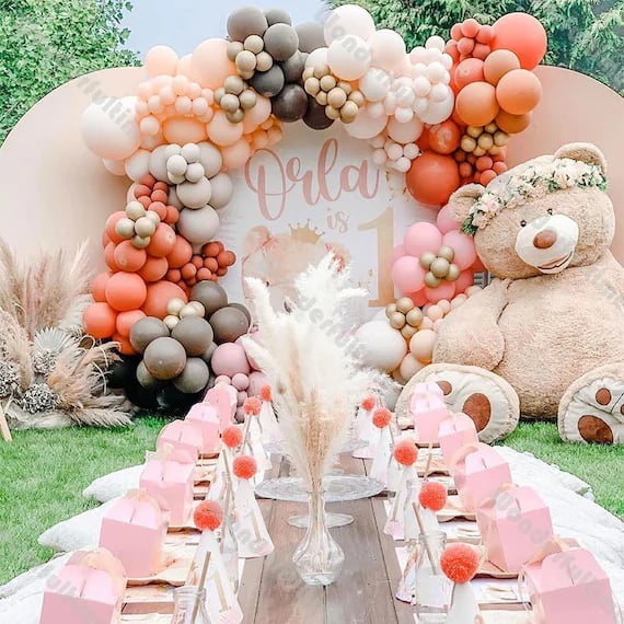 Colorful balloon arch and teddy bear at a 1st birthday party with a decorated table set outdoors.