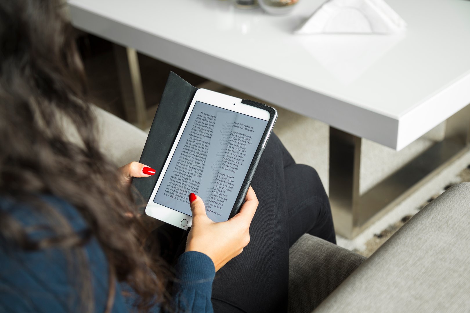 A young woman sitting in the living room on a couch and reading a book on her tablet computer.
