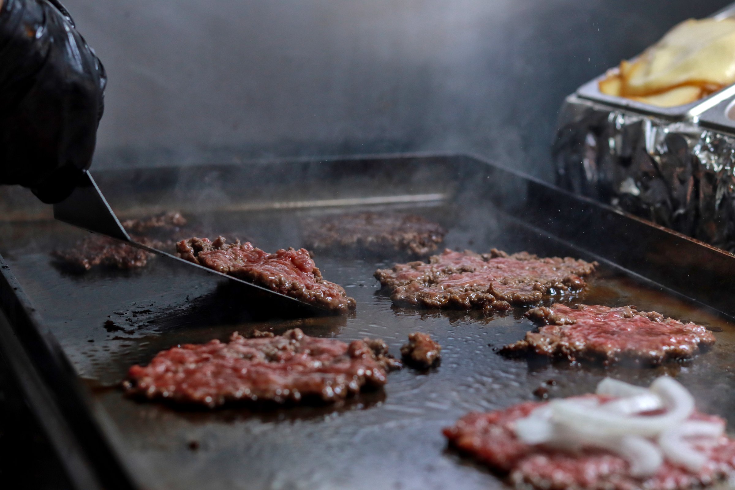 Smash burgers being prepared on a griddle with onions, showing the sizzling cooking process in a professional kitchen