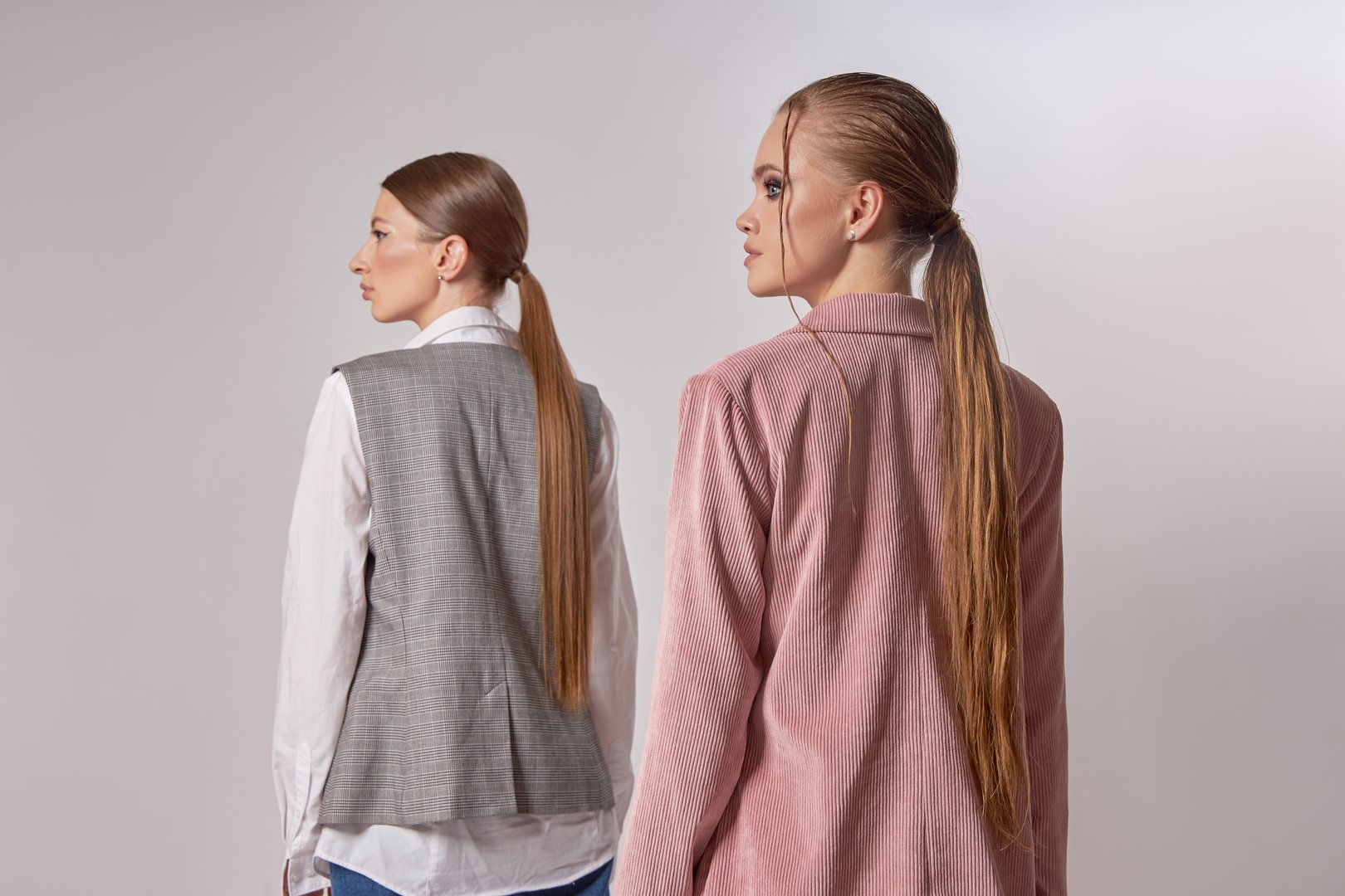 Models pose back-to-back in a studio, dressed in stylish neutral outfits with long hair pulled back. focus is on contemporary fashion and the unique textures of their clothing choices.