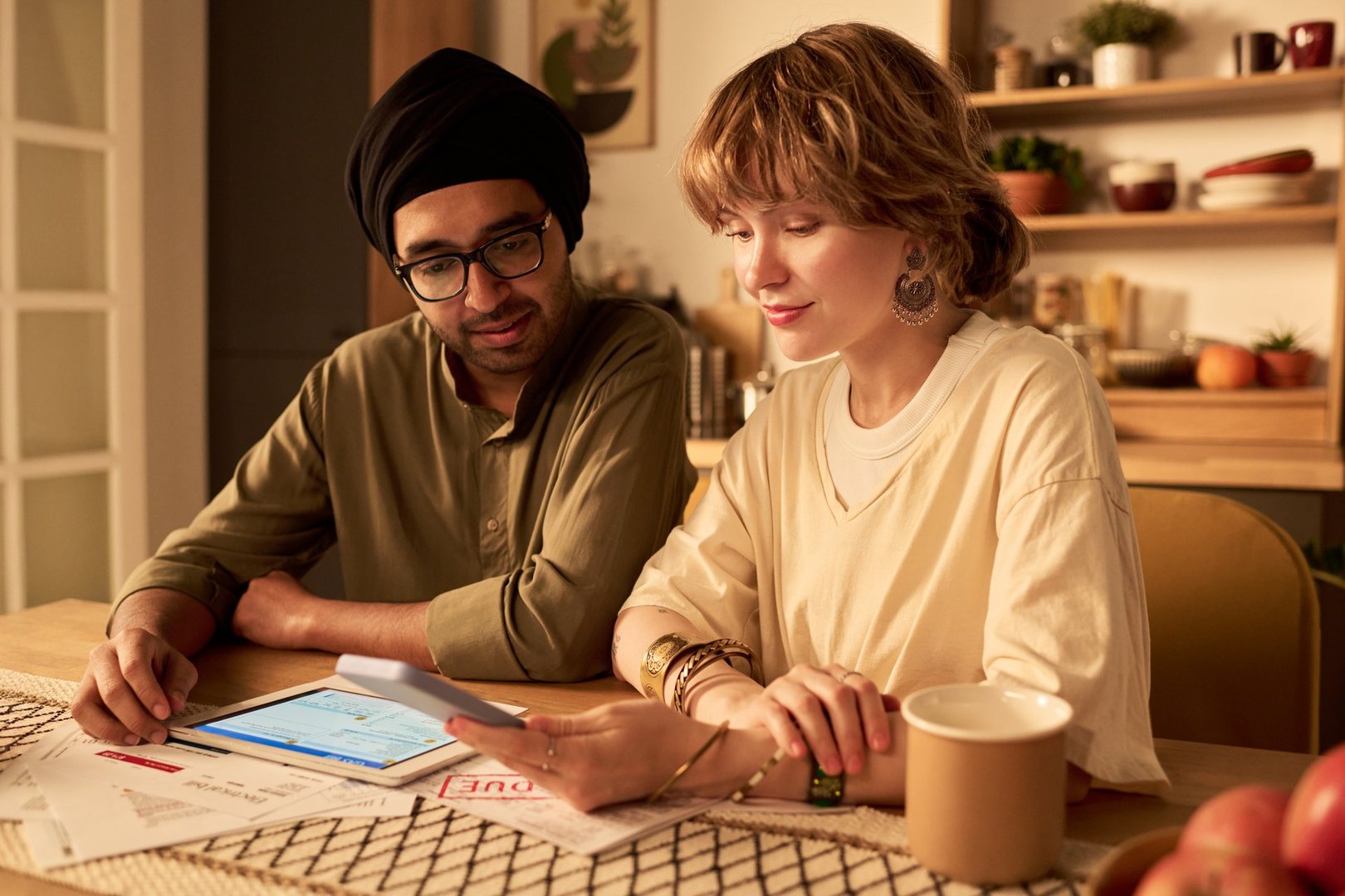 Two individuals collaborating at home office with tablet and documents on table. Plants, coffee mug, and kitchen items visible in background creating a warm ambiance