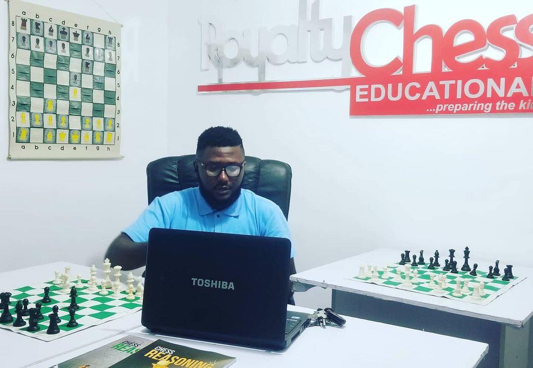 Person sitting at a desk with a laptop, surrounded by chess boards, in a room with a chess-themed poster and logo on the wall.