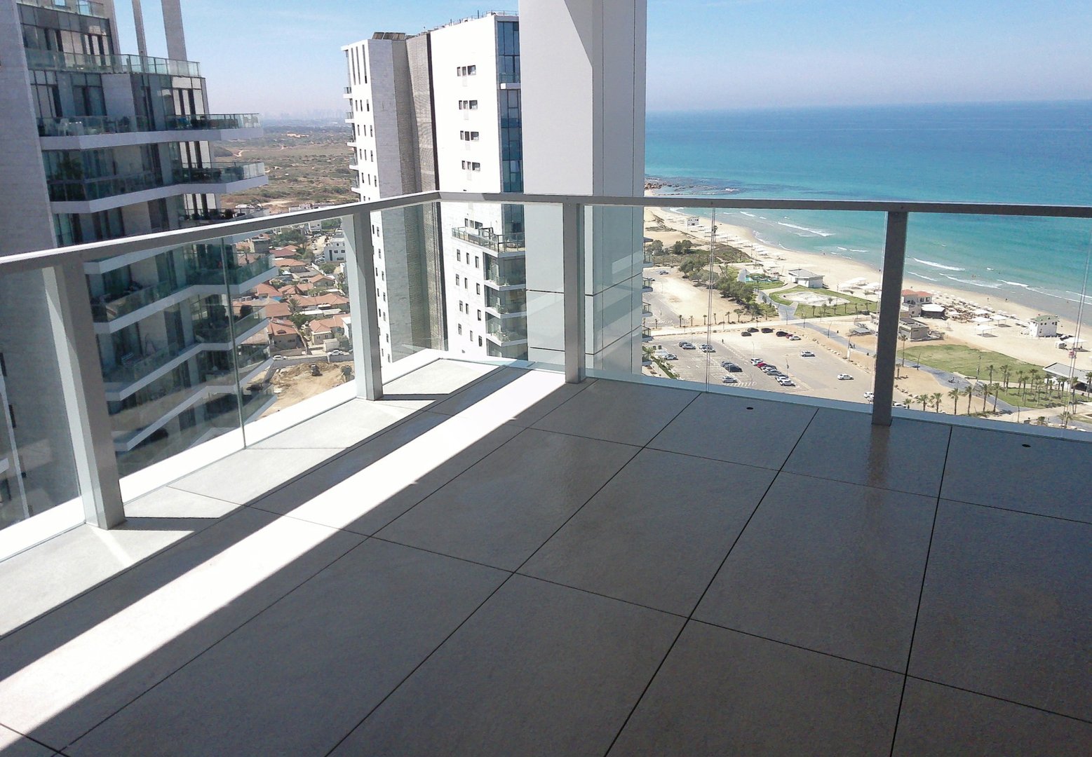 Balcony with glass railing overlooking a beach, ocean, and modern high-rise buildings under a clear blue sky.