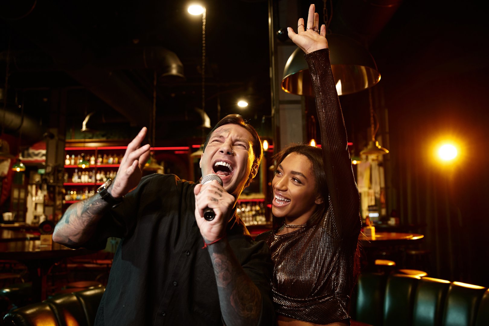 A young multicultural couple shares a joyful moment while singing karaoke at a vibrant bar.