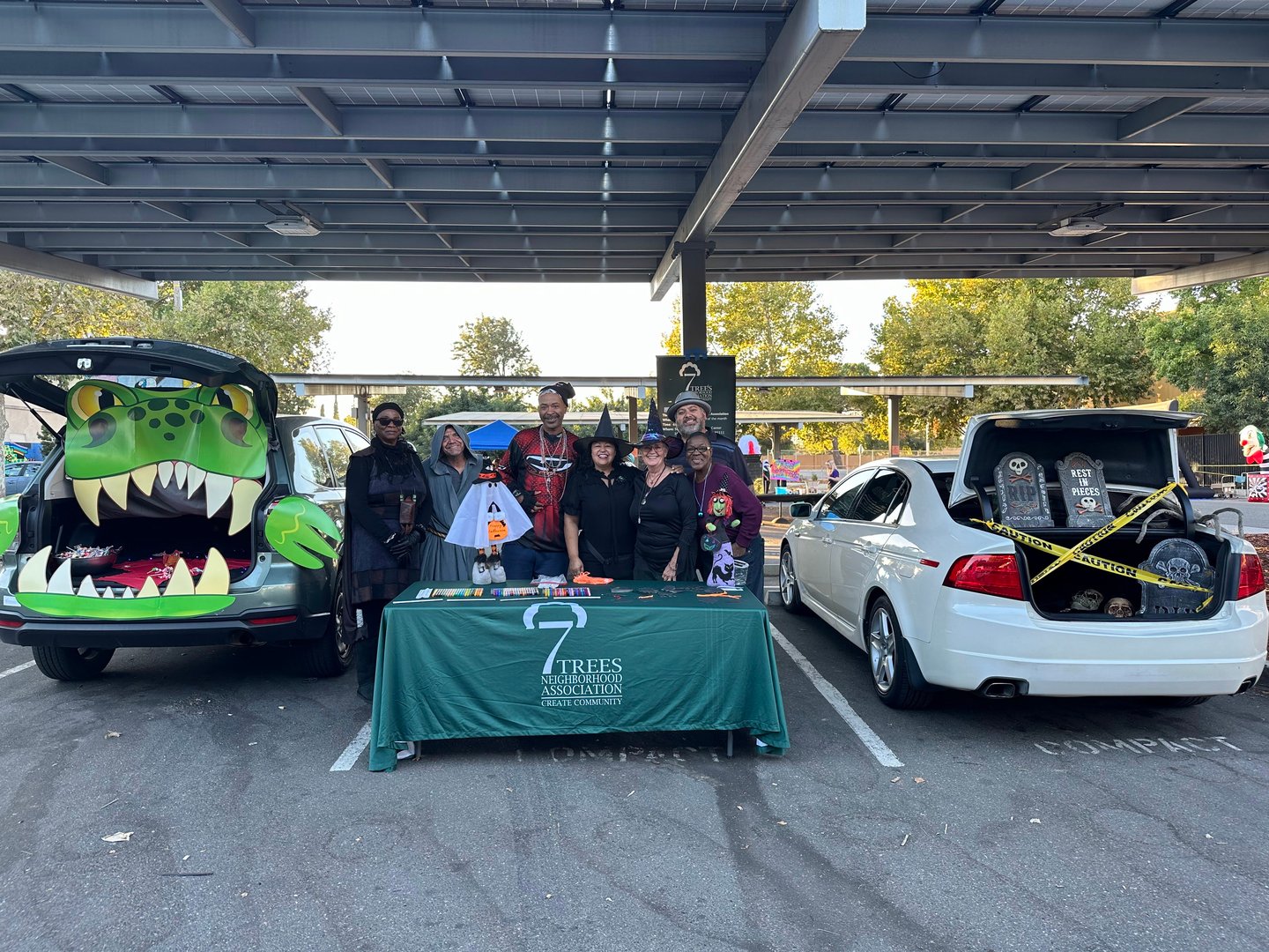 Group dressed in costumes at a booth with Halloween-themed car displays in a parking lot.