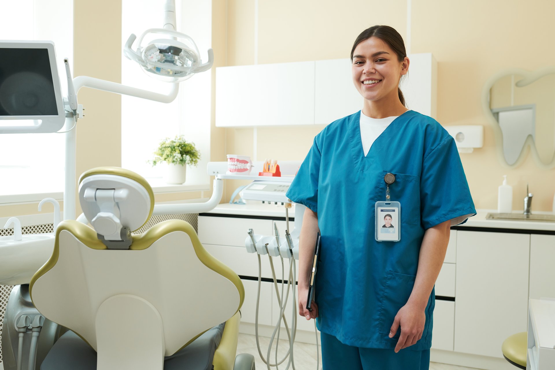 Portrait of young adult Caucasian woman smiling while standing in dental clinic wearing medical scrubs and identification badge, dental chair and equipment visible in background