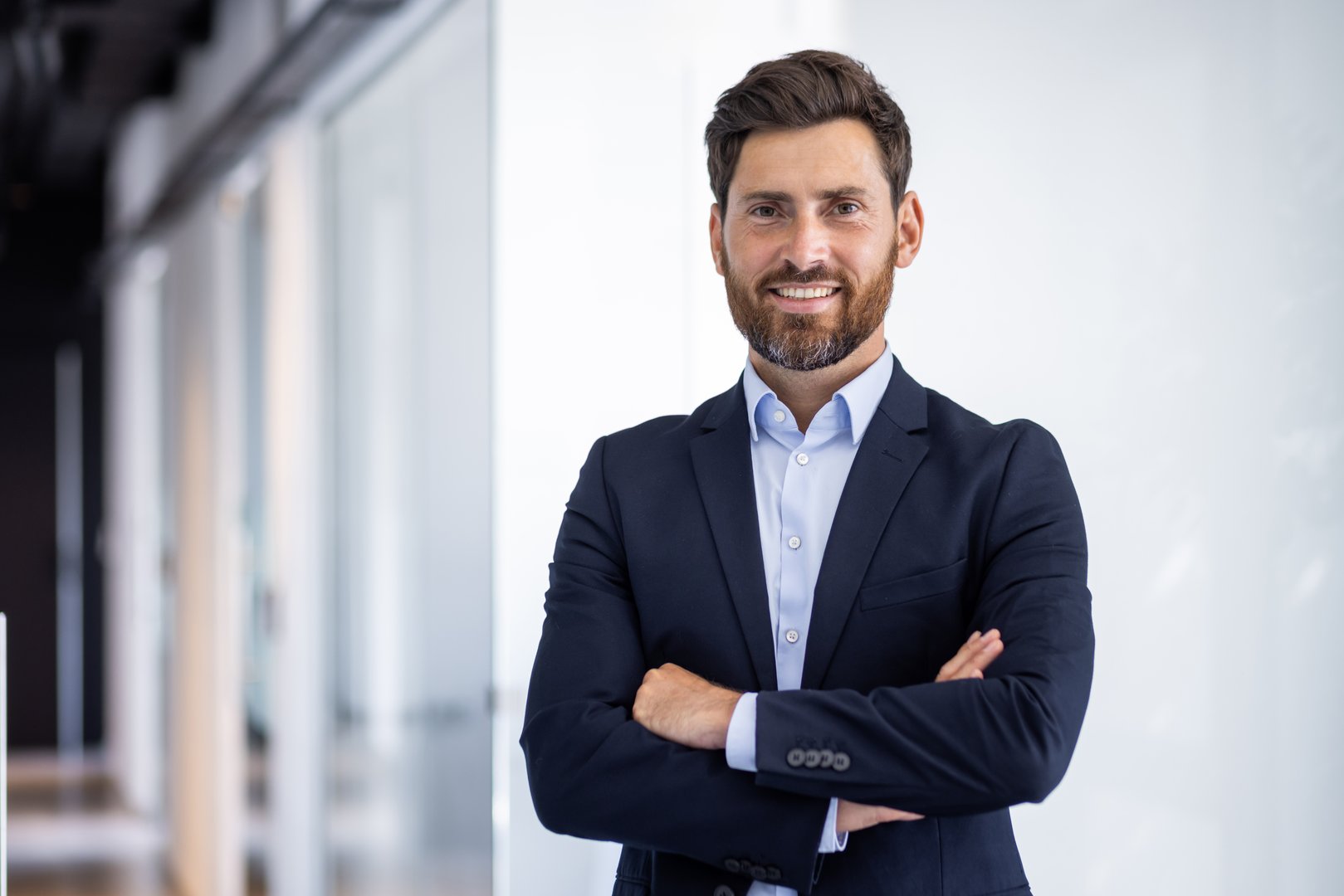 Portrait of a smiling young successful male businessman standing in the office in a suit and looking at the camera with his arms crossed on his chest.