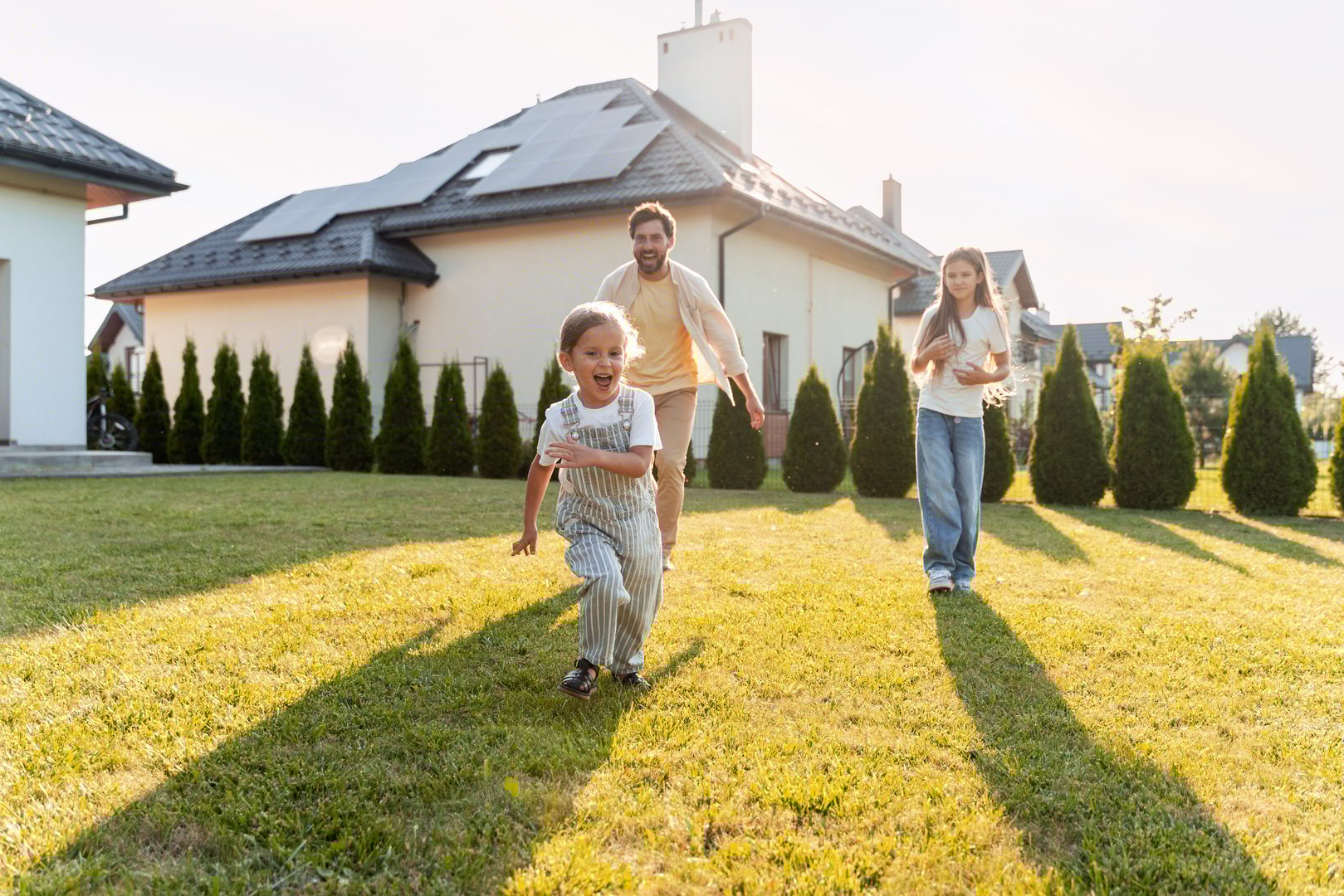 Father and children running on the lawn in front of their house with solar panels on the roof, enjoying a sunny day, symbolizing sustainable living and family fun