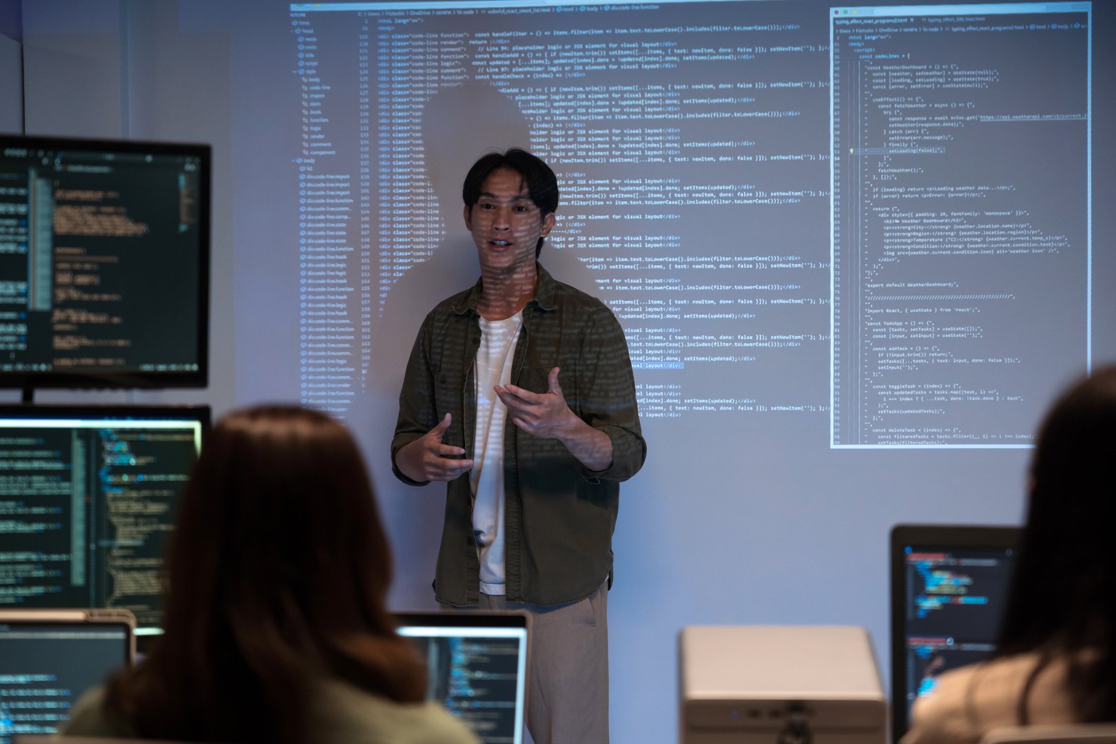 Asian man presents code using a projector during a software development meeting or coding workshop, guiding attendees through problem-solving and programming concepts in an educational setting.