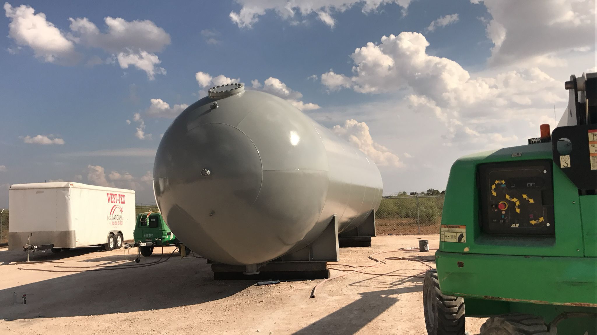 Large metal storage tank on a construction site with equipment and a utility trailer under a blue sky with clouds.