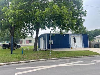 A worker on a ladder painting a blue building with white doors under a tree on a grassy roadside.