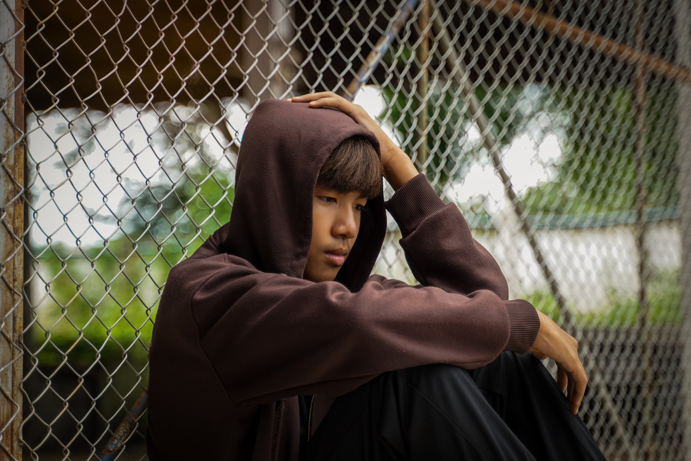 Asian boy wearing a hoodie sits with his knees bent, his back leaning against a wire mesh fence, his face in his hands, his head in his hands. The concept of troubled teenagers, loneliness, depression.