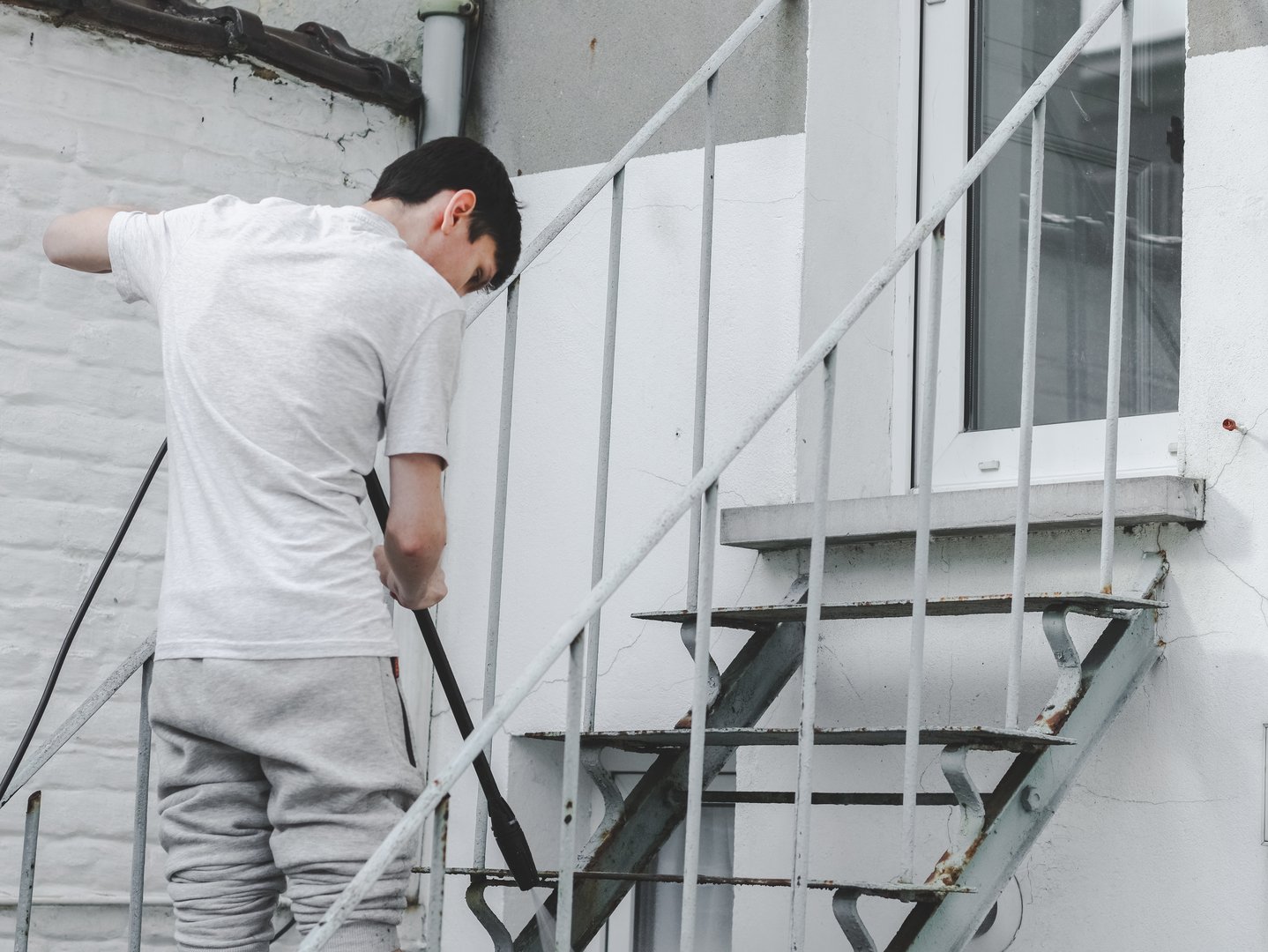 Caucasian guy in gray sportswear and a t-shirt on the side washes with a karcher with water under pressure the steps of a metal ladder up in the backyard of his house, side view close-up.The concept of washing the yard,holiday preparation,at home,cleaning services.