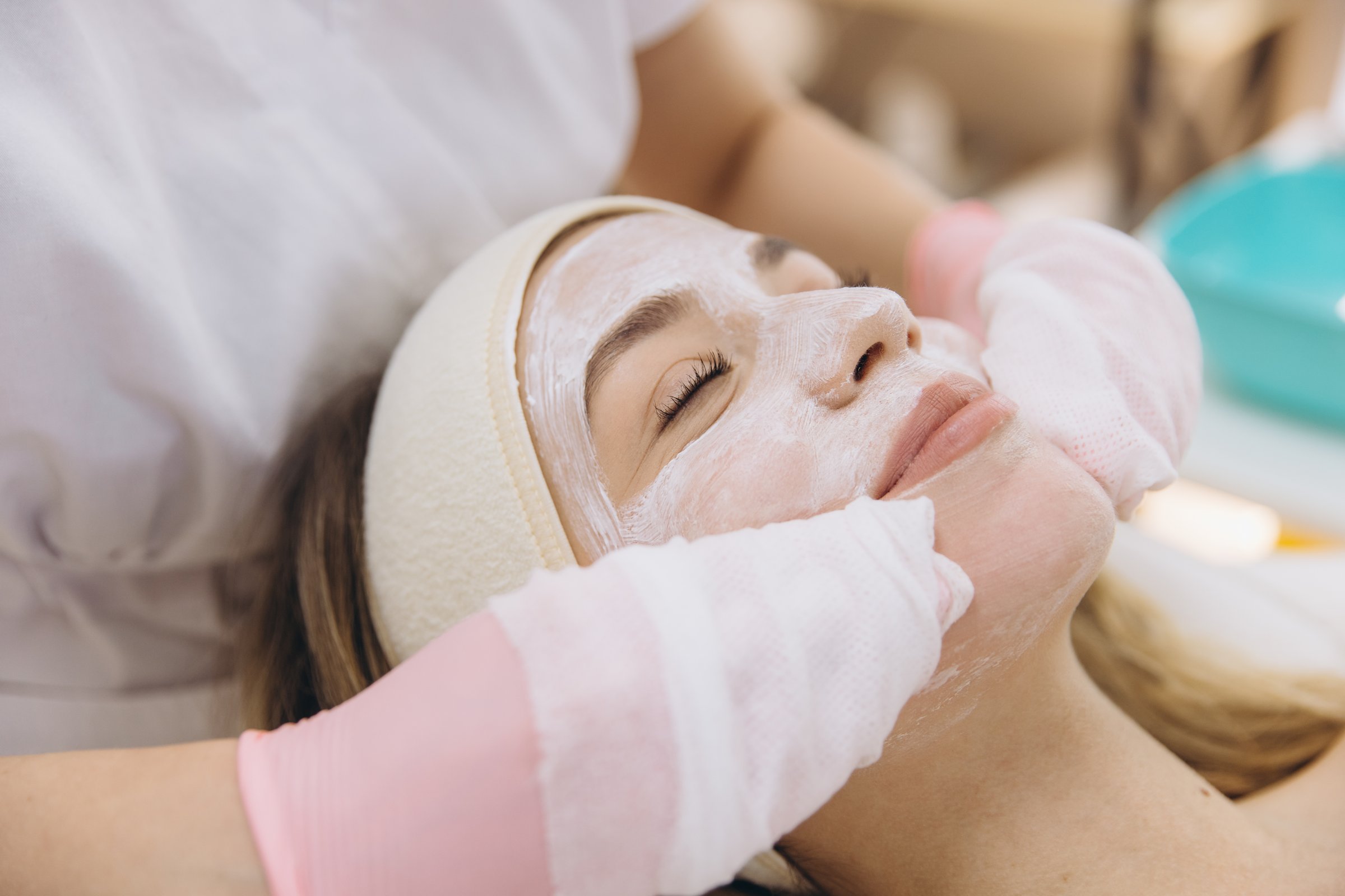 Gloved beautician spreading white facial mask across client's skin during professional skincare treatment in spa setting