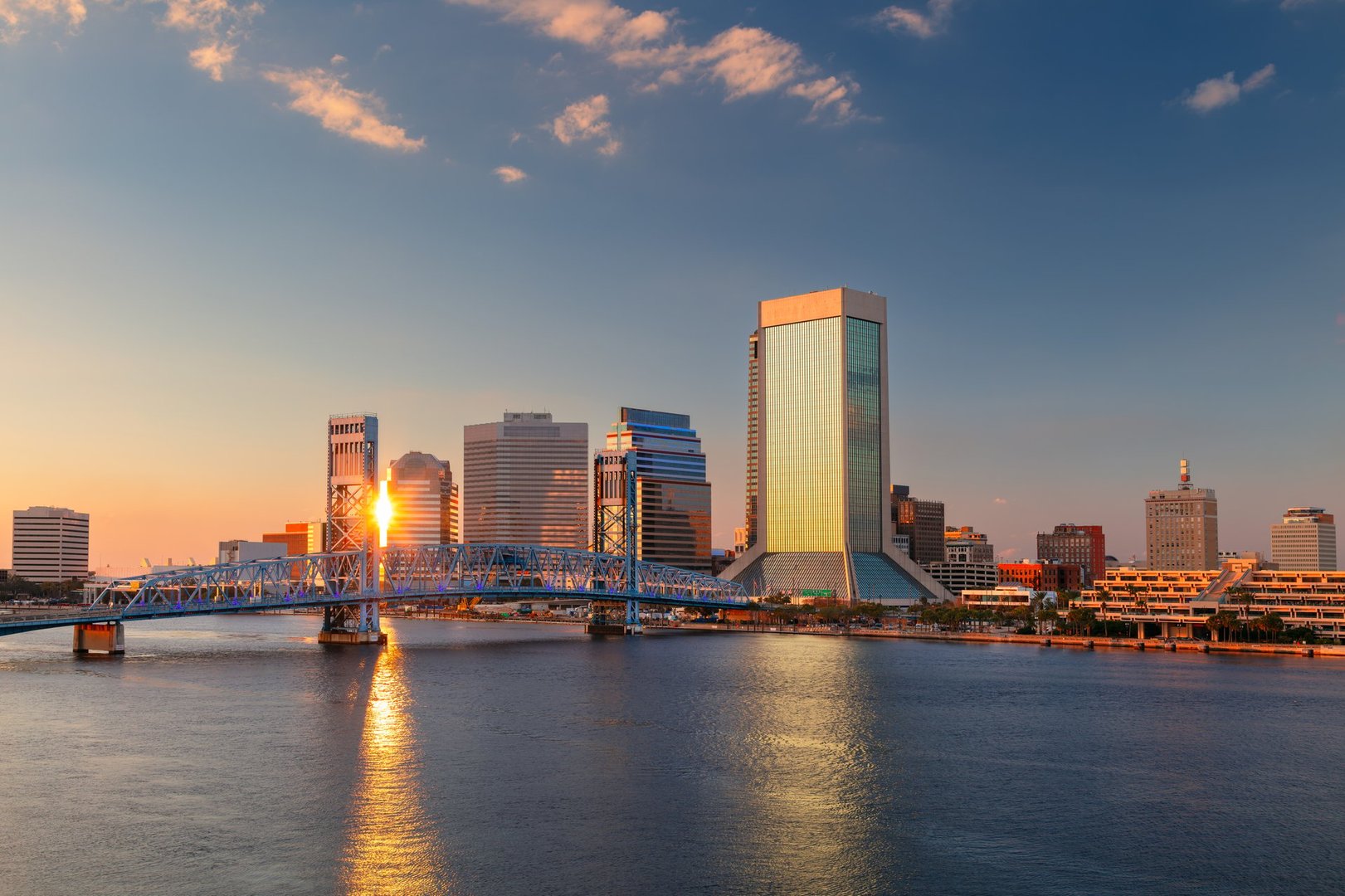 Skyline of Jacksonville, Florida, featuring a mix of modern skyscrapers reflecting golden sunset hues. The prominent blue John T. Alsop Jr. Bridge spans the St. Johns River, connecting both sides. Warm light bathes the buildings, highlighting glass facades and diverse architecture. Sparse clouds accentuate the clear evening sky, creating a tranquil scene. The river reflects the vibrant colors, adding to the city's picturesque charm at dusk.