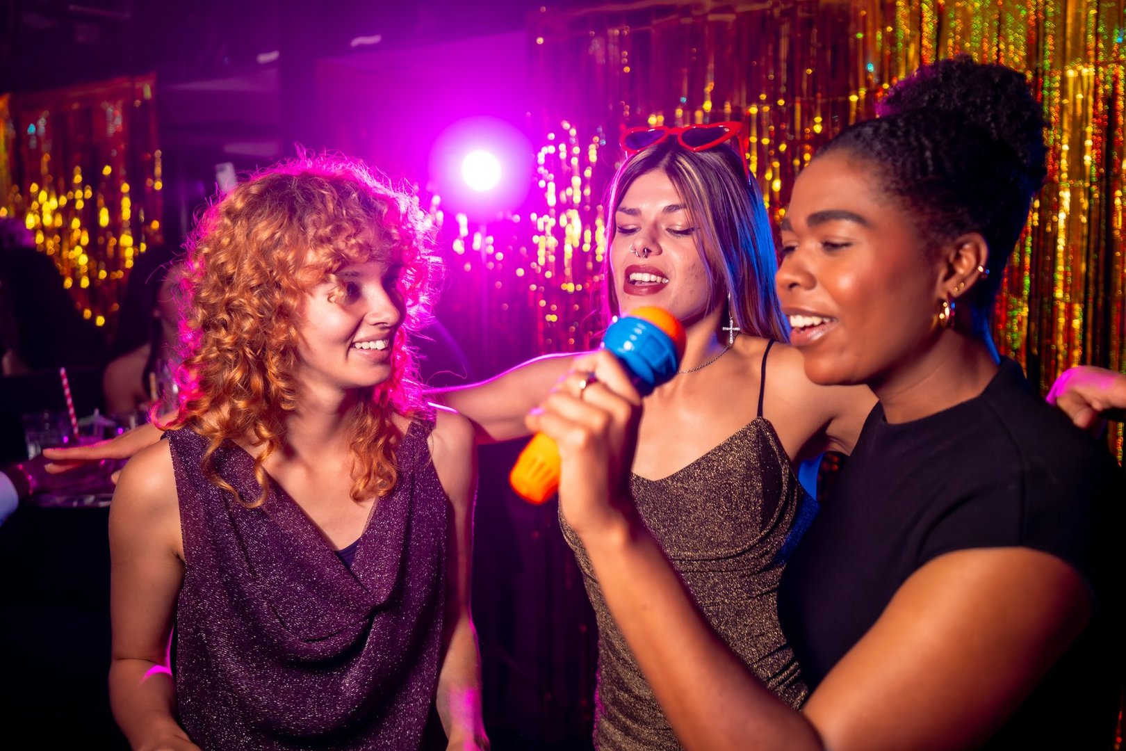 Three multi-ethnic young women enjoying a lively karaoke night at a trendy nightclub, singing together and sharing joyful moments