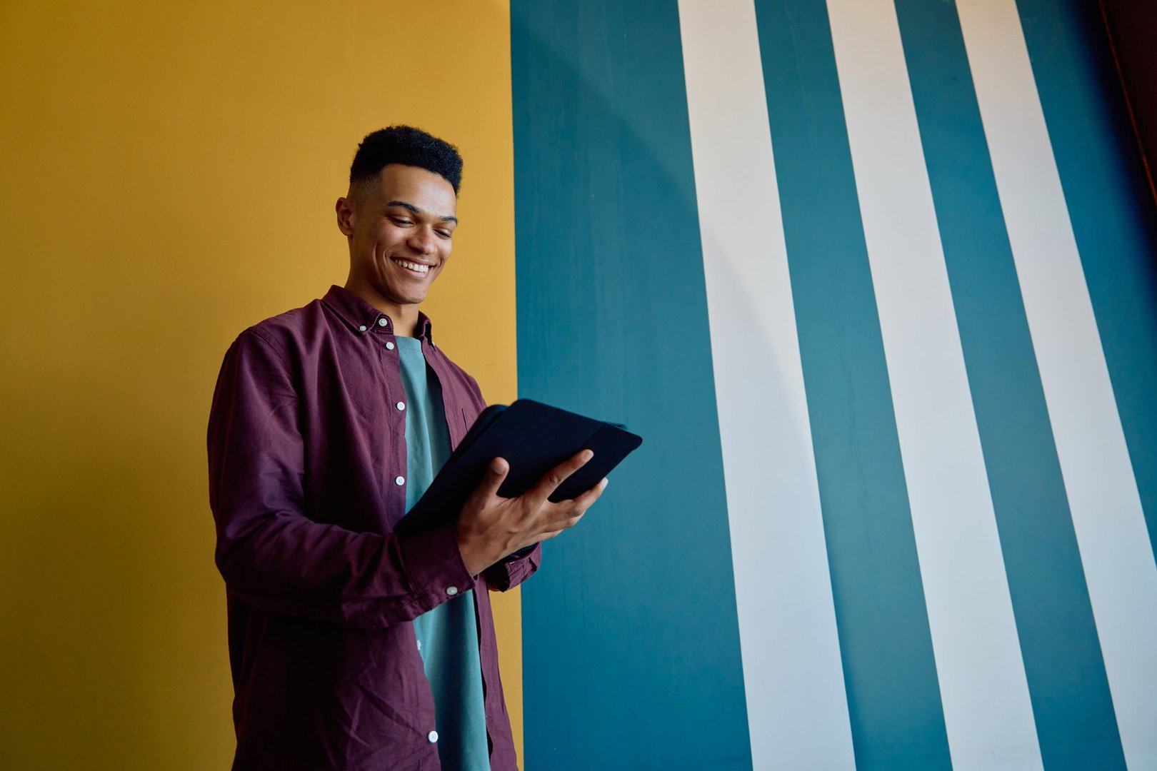 Smiling young businessman in smart casual using tablet while standing against colorful wall in modern office