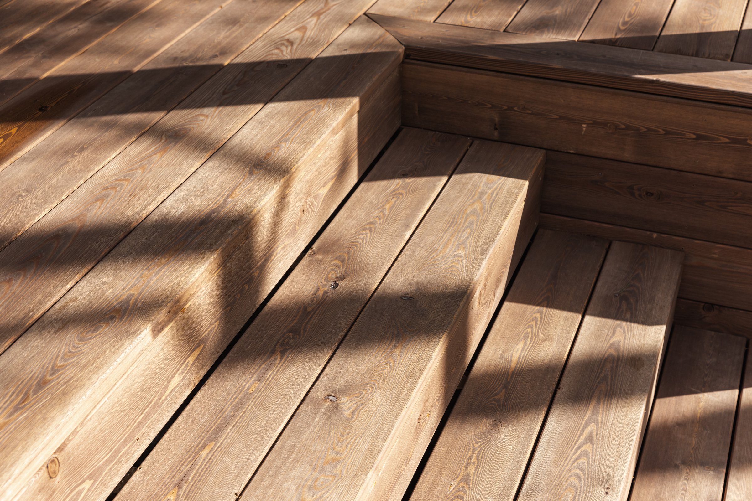 Wooden deck stairs are captured in soft natural daylight, showcasing their grain and texture. The shadows cast create an interesting interplay of shapes, adding depth to the scene
