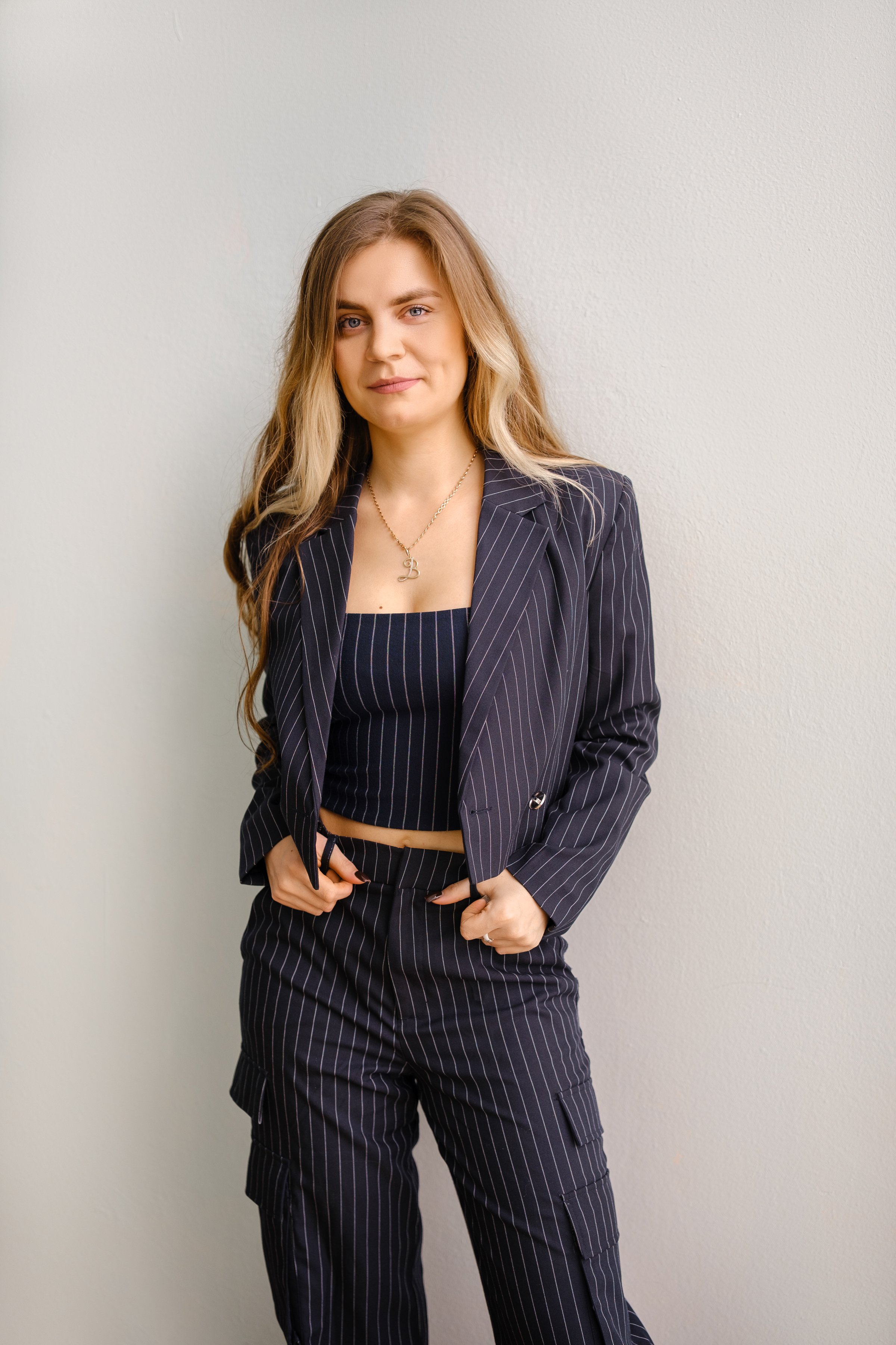 A young woman stands against a plain wall, exuding confidence in a fitted striped suit. Her stylish outfit features a cropped top and tailored pants, showcasing a modern and chic aesthetic.