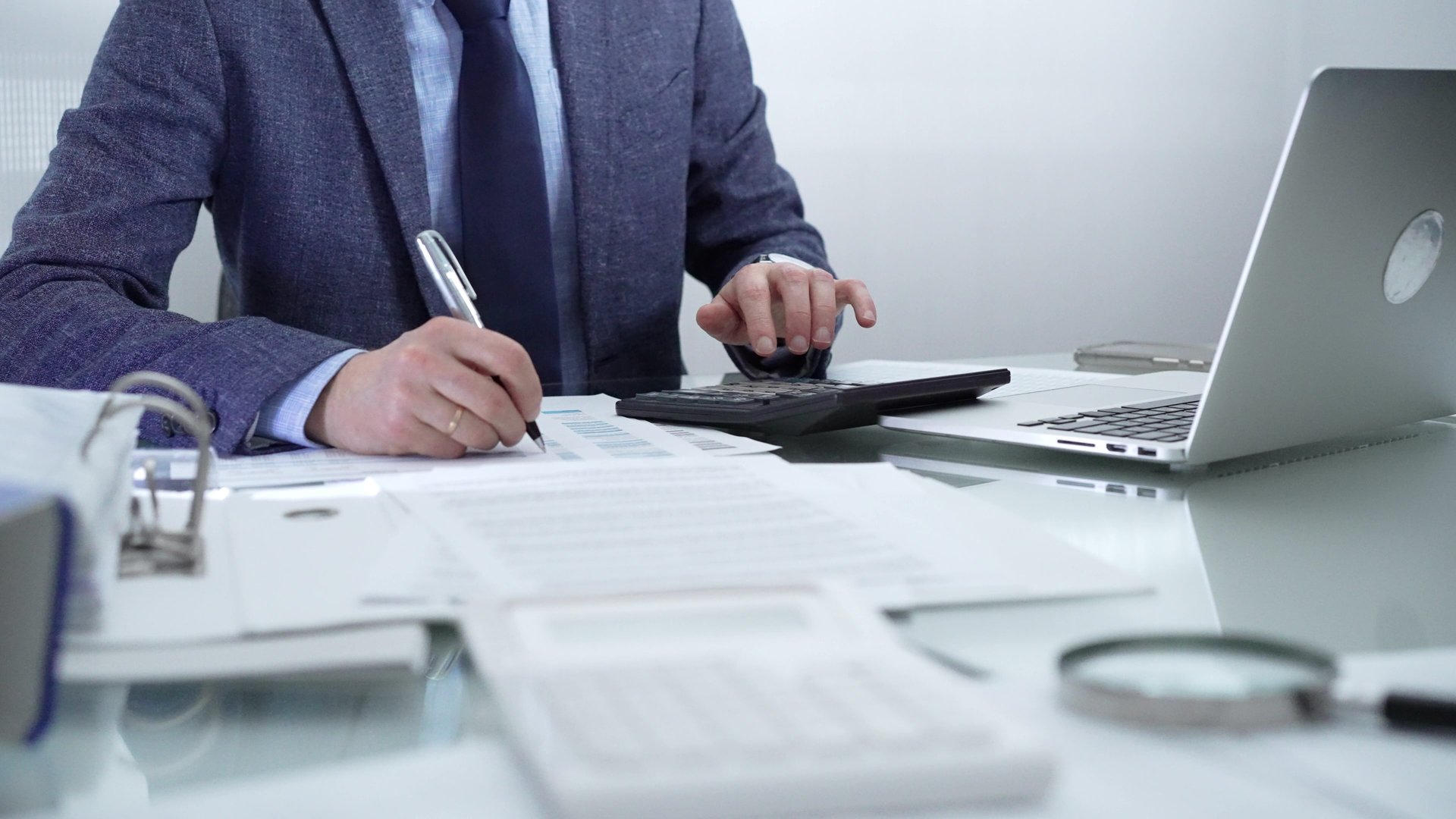 Businessman wearing blue suit is working with laptop, calculator and documents in a professional office setting. Low lighting, close-up view. Business people, audit concept.