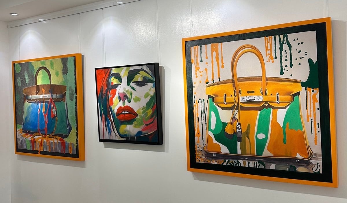 Three colorful paintings on a white wall; two feature handbags and one abstract portrait with bold colors.