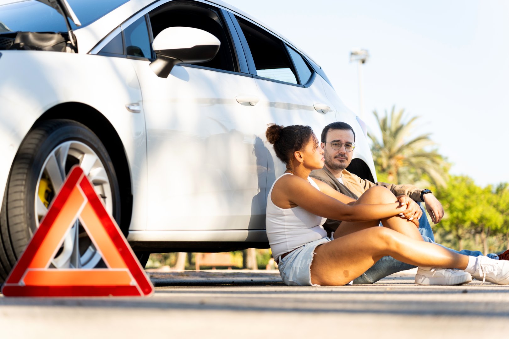 Young multi-ethnic couple sitting on the asphalt by their broken-down car with open hood and warning triangle, waiting for help
