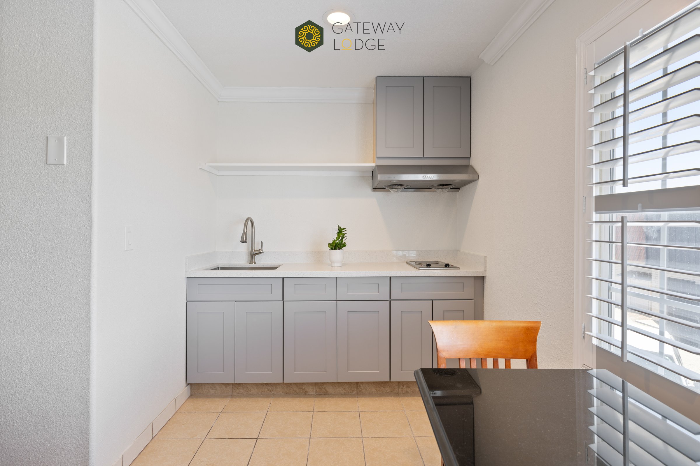 Small kitchenette with gray cabinets, stove, sink, and a plant. Wall sign reads Gateway Lodge. Wooden chair and shutters visible.