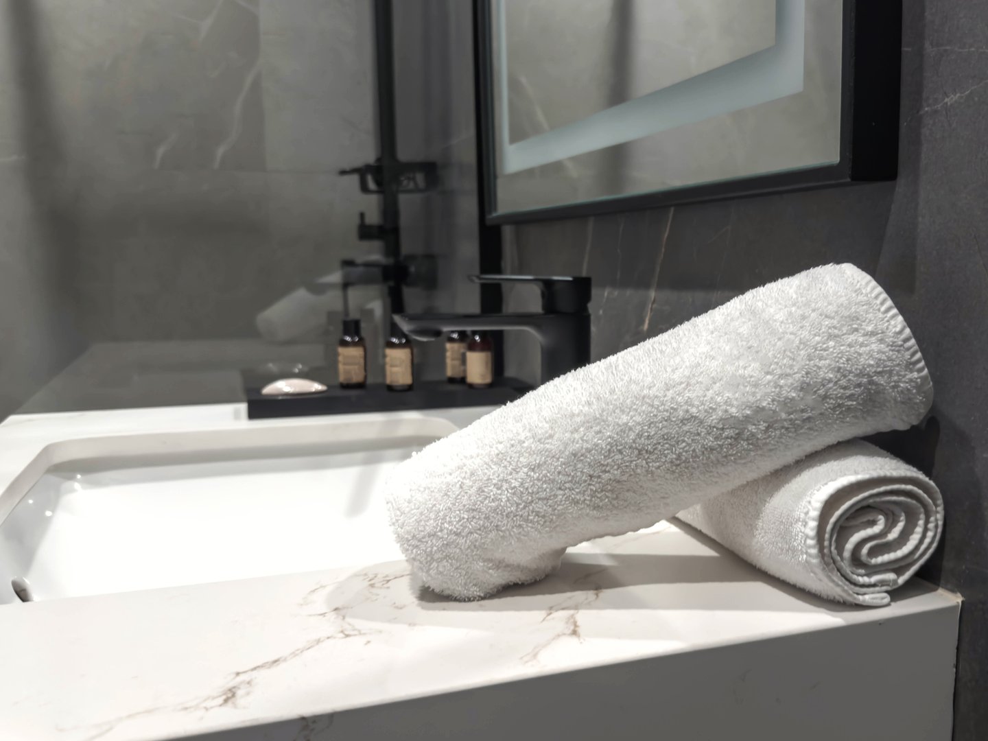Modern hotel bathroom with soft white towels neatly rolled on a marble countertop near a stylish black faucet and amenities, creating a clean, luxurious and relaxing spa-like atmosphere for guests.