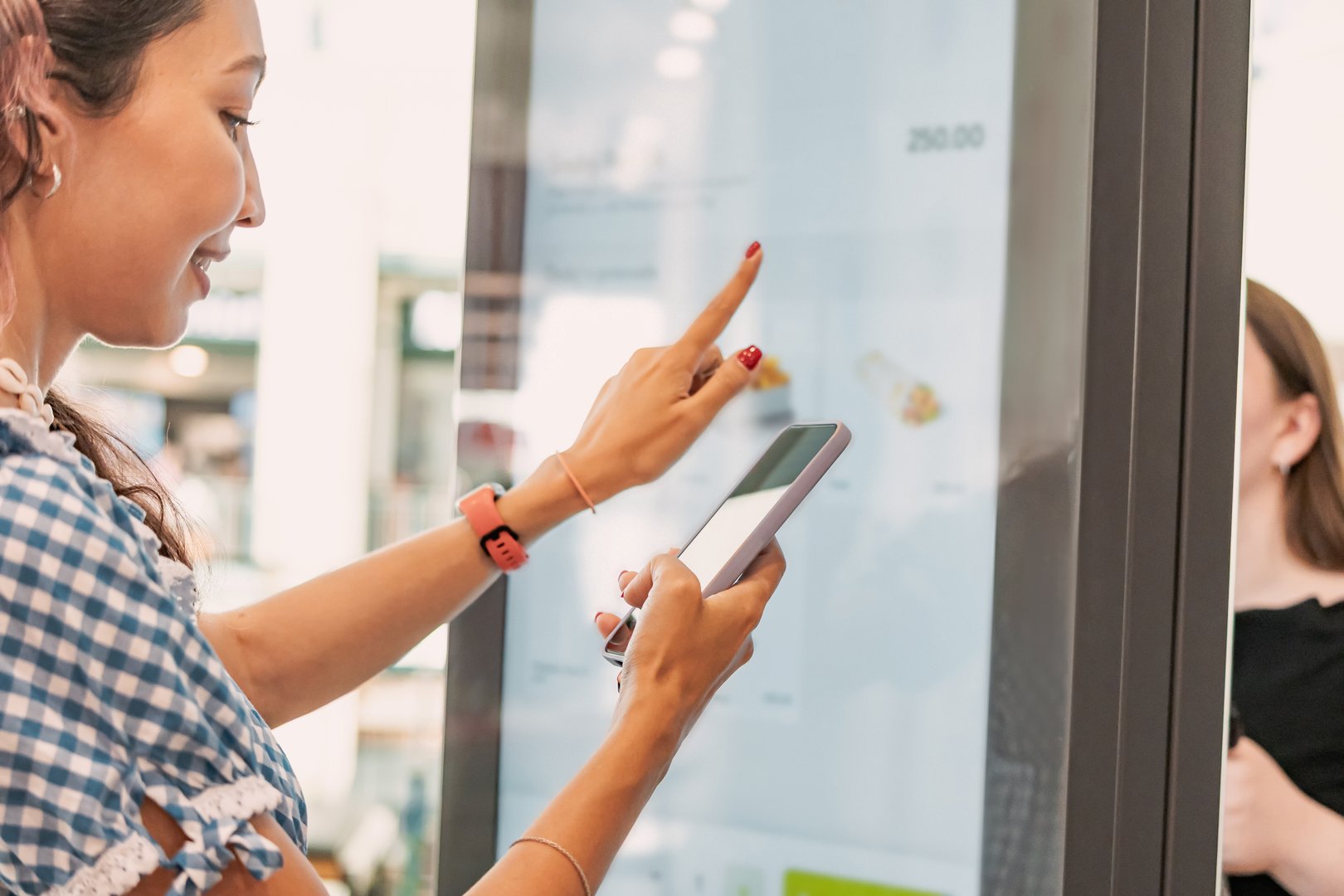 Customer using smartphone and interactive self ordering kiosk at fast food restaurant to choose meal and make contactless payment
