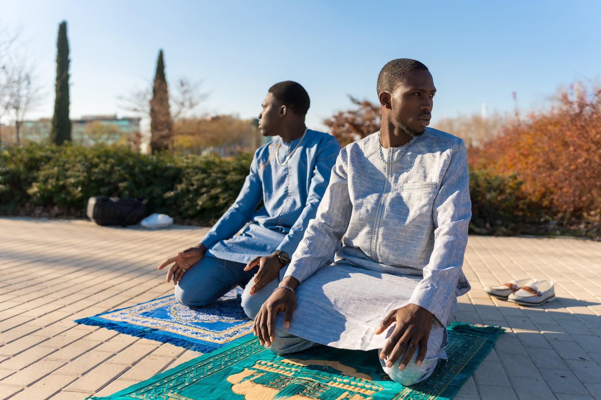 Two young african muslim men are kneeling on prayer rugs outdoors, praying in traditional dashiki clothing