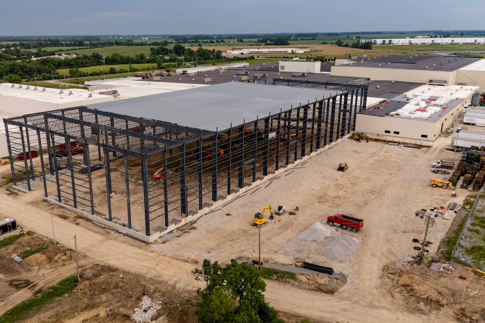 The image shows a vast construction site, featuring the skeletal structure of a future warehouse or factory surrounded by open fields and distant structures.