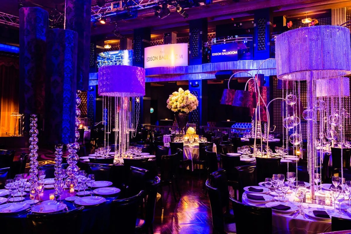 Elegant ballroom with ornate chandeliers, round tables set for an event, and vibrant purple and blue lighting.