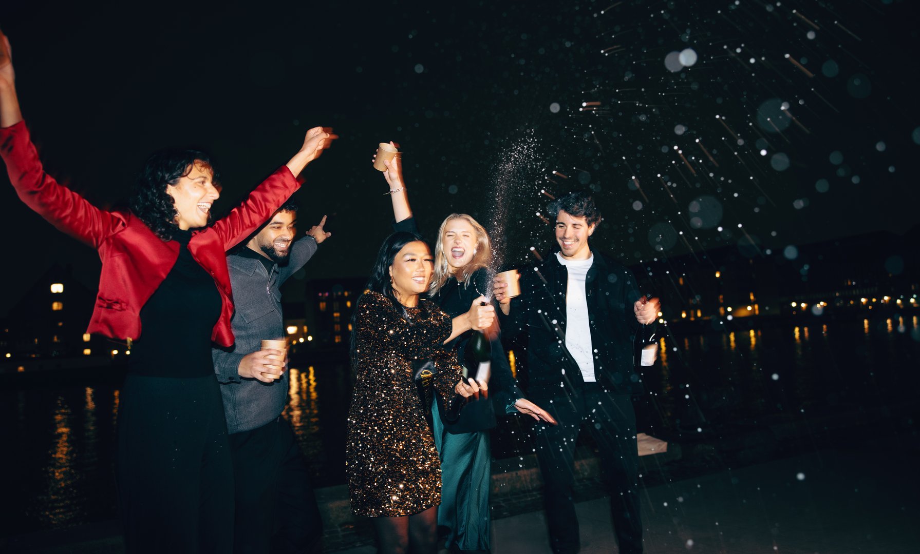 A group of joyful friends celebrates with champagne bottles and sparklers outdoors at night, sharing laughter and excitement amidst sparkling fireworks over a stunning cityscape.